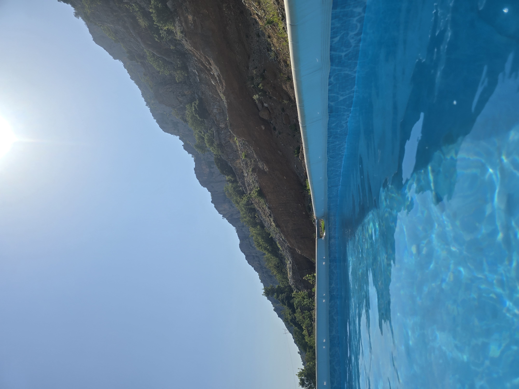 In an outdoor setting, a clear blue swimming pool occupies the foreground, its surface rippling gently. Beyond the pool, a steep, rocky mountainside dominates the midground and background, dotted with sparse green vegetation. The sky above is a brilliant, cloudless blue, with the sun casting a bright glare near the top center of the image. The scene suggests a natural, rugged environment adjacent to a man-made amenity. The location is identified as Unknown, Unknown.