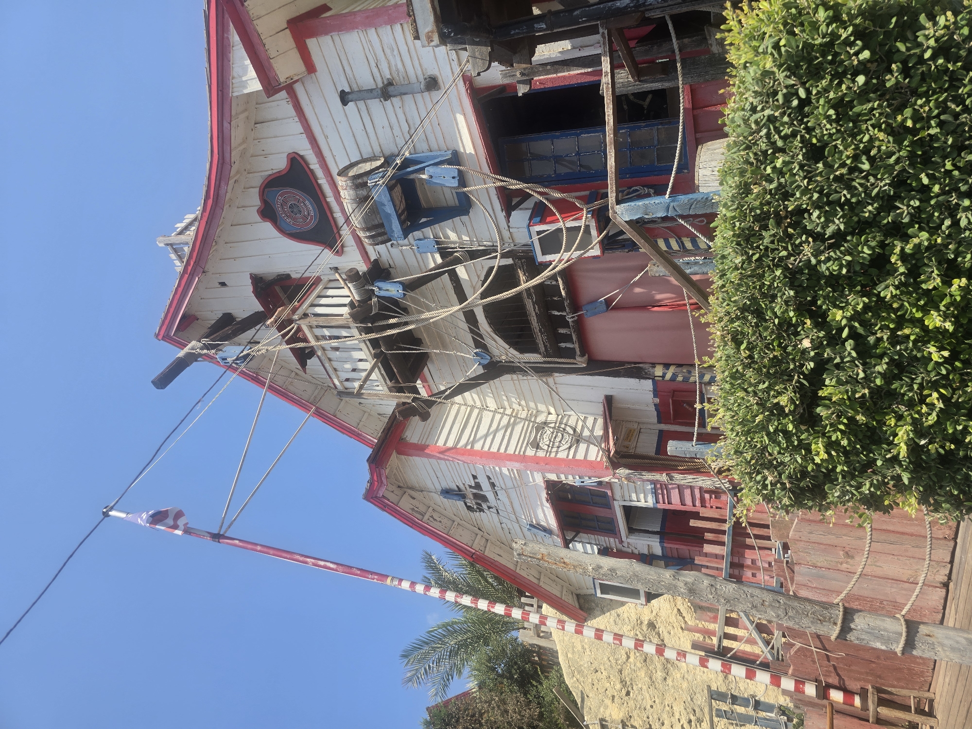 The image shows the Popeye Village, a film set and tourist attraction located in the town of Mellieħa, Malta. The village is characterized by its collection of colorful, rustic wooden buildings designed to resemble a quaint fishing village. In the foreground, a well-maintained green hedge partially obscures the view of the buildings. A wooden fence with red and white stripes lines the left side of the image, adjacent to a sandy embankment with some greenery. A wooden pole with red and white stripes stands prominently on the left, with ropes attached to it. A flag, which appears to be the flag of Malta, flies from a flagpole positioned on top of one of the buildings. The buildings themselves are constructed with white wooden planks and adorned with red and blue trim. Various nautical elements are visible, including ropes, pulleys, a barrel, and what appears to be a ship's wheel through an open doorway. The sky above is a clear, bright blue, indicating a sunny day. The overall scene evokes a whimsical and playful atmosphere, typical of a theme park or entertainment venue.