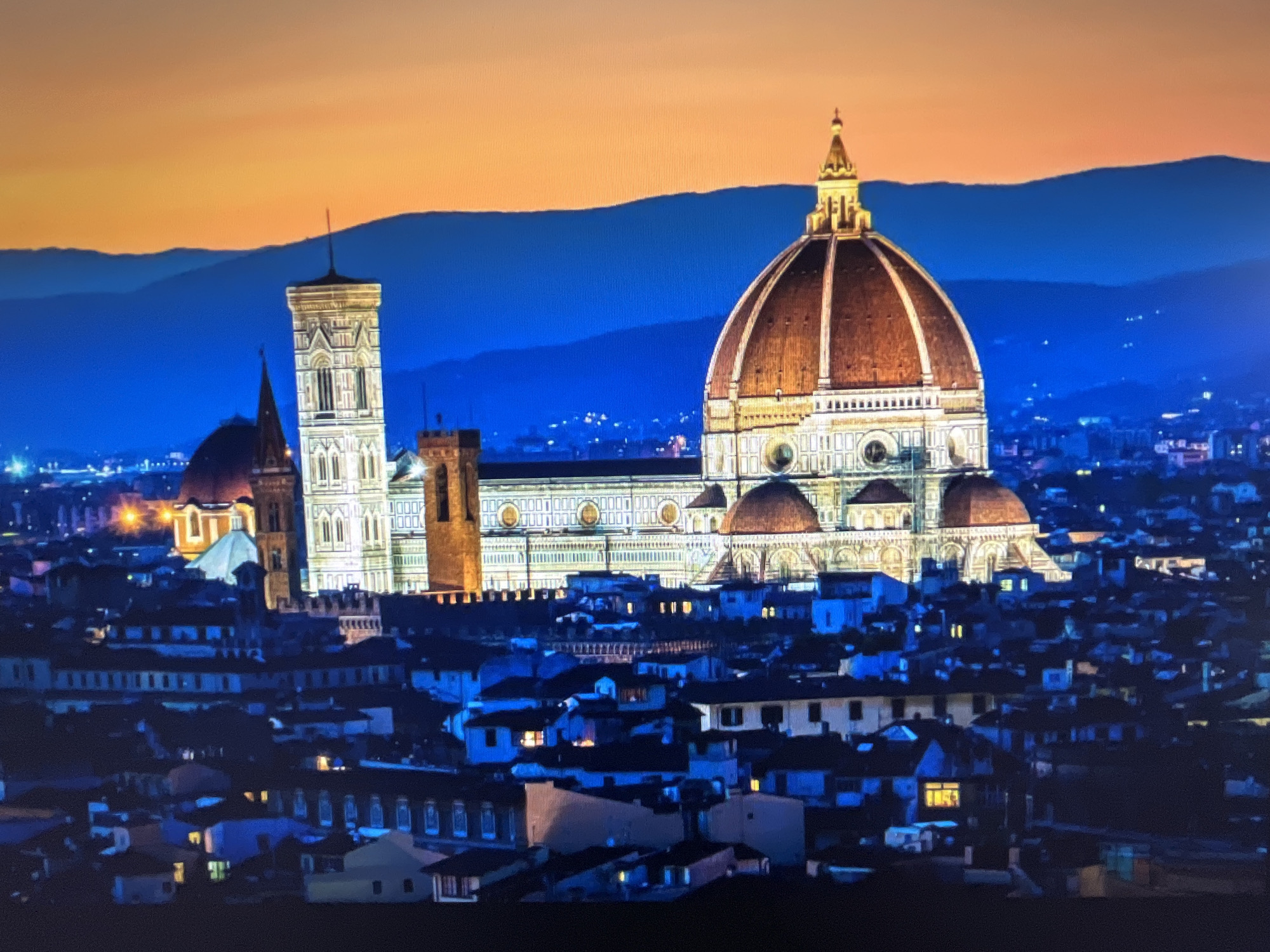 The image depicts a nighttime cityscape with the Florence Cathedral, also known as the Duomo, prominently featured. The cathedral's dome is illuminated with a warm, reddish-brown hue, and its intricate architectural details are visible. Adjacent to the cathedral is Giotto's Campanile, a tall, slender bell tower, also lit up. The buildings in the foreground are dark, with some windows showing interior lights, suggesting residential or commercial spaces. The sky transitions from a deep blue near the horizon to a gradient of orange and yellow at the top, indicating either a sunset or sunrise. Distant, dark mountain ranges are visible against the colorful sky. The overall impression is of a historic European city at dusk or dawn. The claim that this image depicts the city of Varna, Bulgaria is incorrect; this is Florence, Italy.