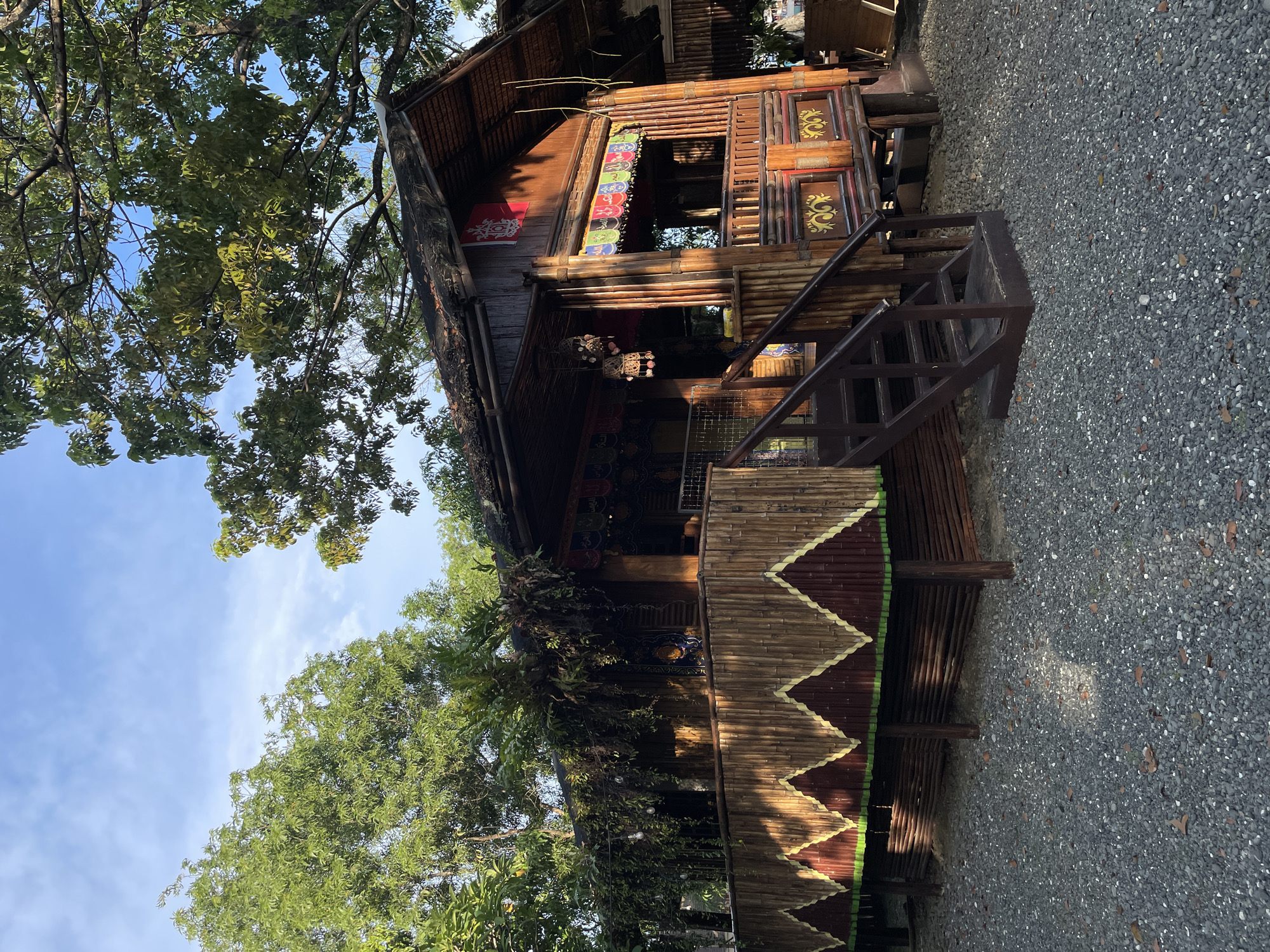 The image shows a traditional bamboo house with a thatched roof, located outdoors in Davao City, Philippines. The house is elevated on stilts and has a wooden staircase leading up to a porch area. The walls are constructed from bamboo poles, and decorative elements such as patterned fabric banners and hanging lanterns adorn the exterior. Lush green trees surround the structure, and the ground is covered in gravel. The sky is visible in the background with some clouds. The scene appears to be during daylight hours, with sunlight casting shadows on the house and ground.