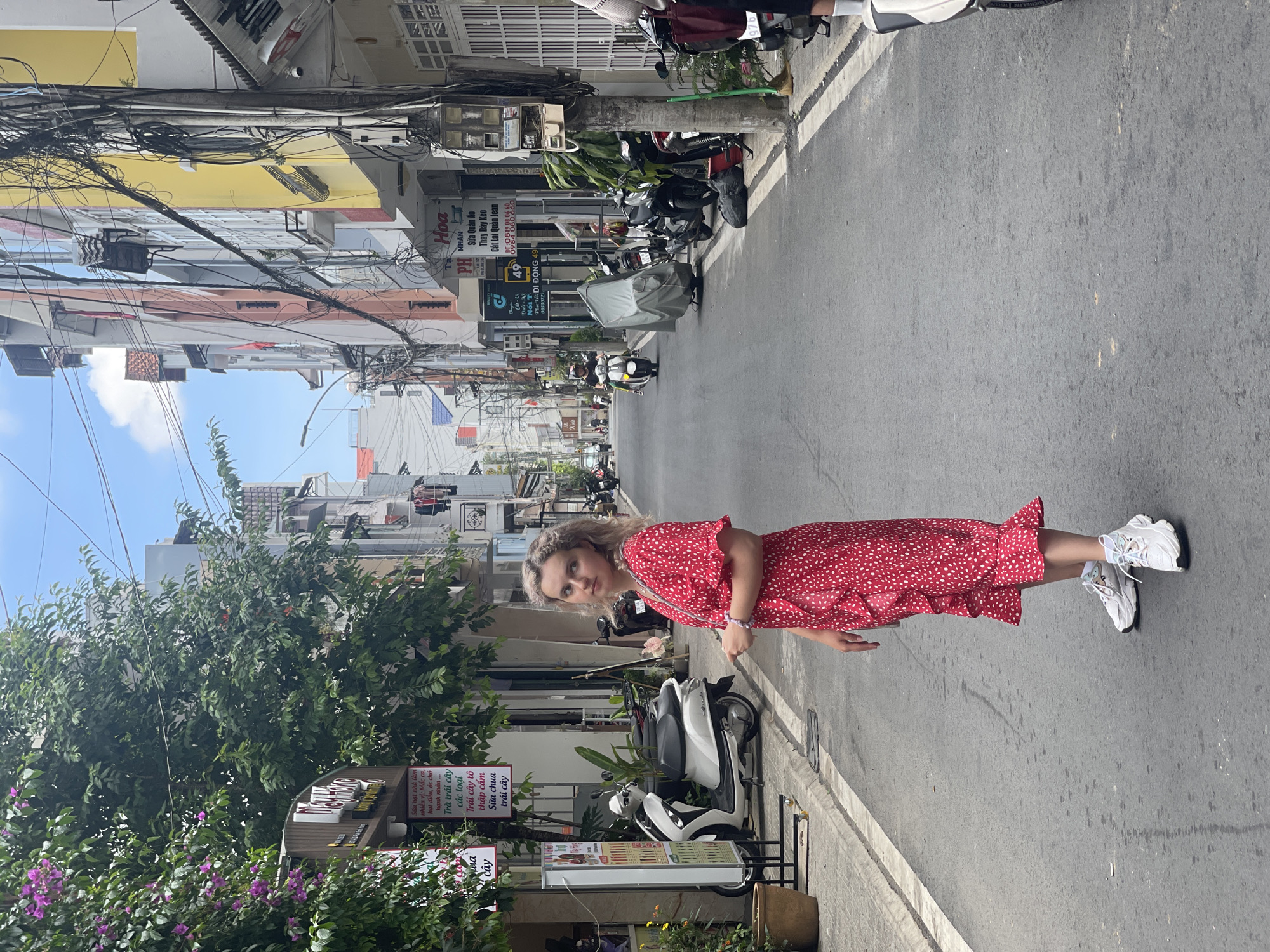 The photograph is of a Caucasian woman standing on a paved street in Da Lat, Vietnam. She is wearing a red dress with white polka dots and white sneakers. The street is lined with buildings, some of which have signs in Vietnamese. There are power lines overhead. There is a scooter parked on the sidewalk in the foreground and other scooters farther down the street. There are signs on the left for a store or restaurant selling produce. Some of the signs have English translations.