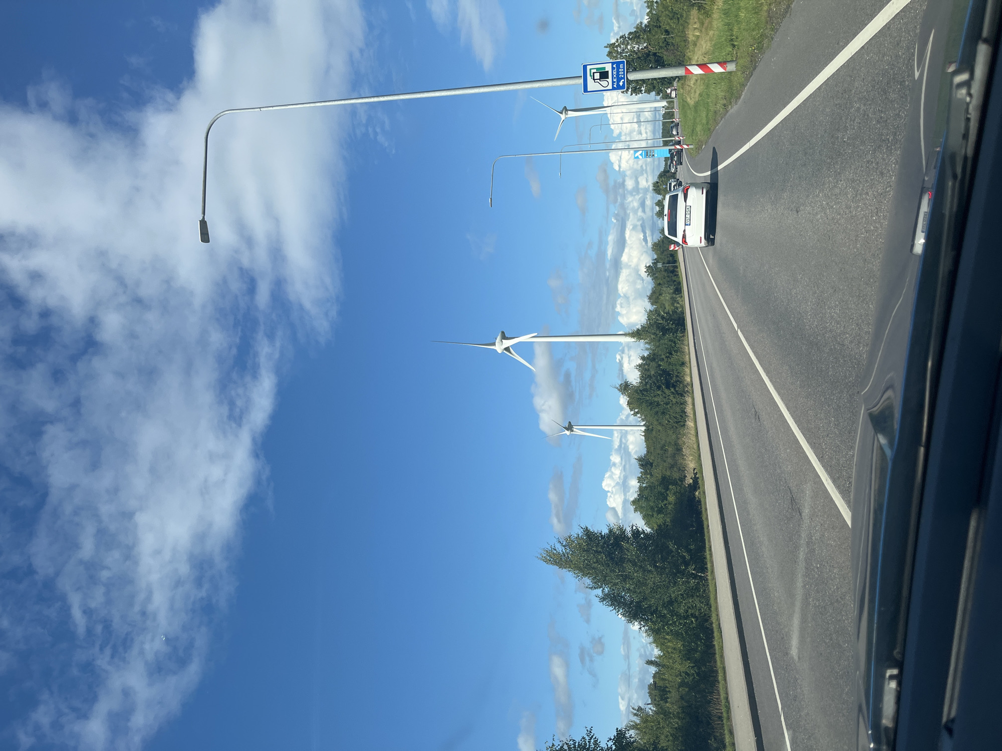 This image, captured on a sunny day in Virtsu, Estonia, depicts a view from a vehicle driving on a road. The sky is a vibrant blue, with scattered white clouds. The road is paved and has multiple lanes marked by white lines. On the right side of the road, there is a green grassy verge and a series of tall lampposts. A blue and white sign indicating "ALEXELA 200m" with a fuel pump icon is also visible on the right side. In the distance, several large white wind turbines are erected, suggesting a wind farm. A white car is in the right lane ahead, and other vehicles are visible further down the road. Trees and other vegetation line the left side of the road. The front of the vehicle from which the photo is taken is visible at the bottom of the frame, with the dashboard and windshield wipers.