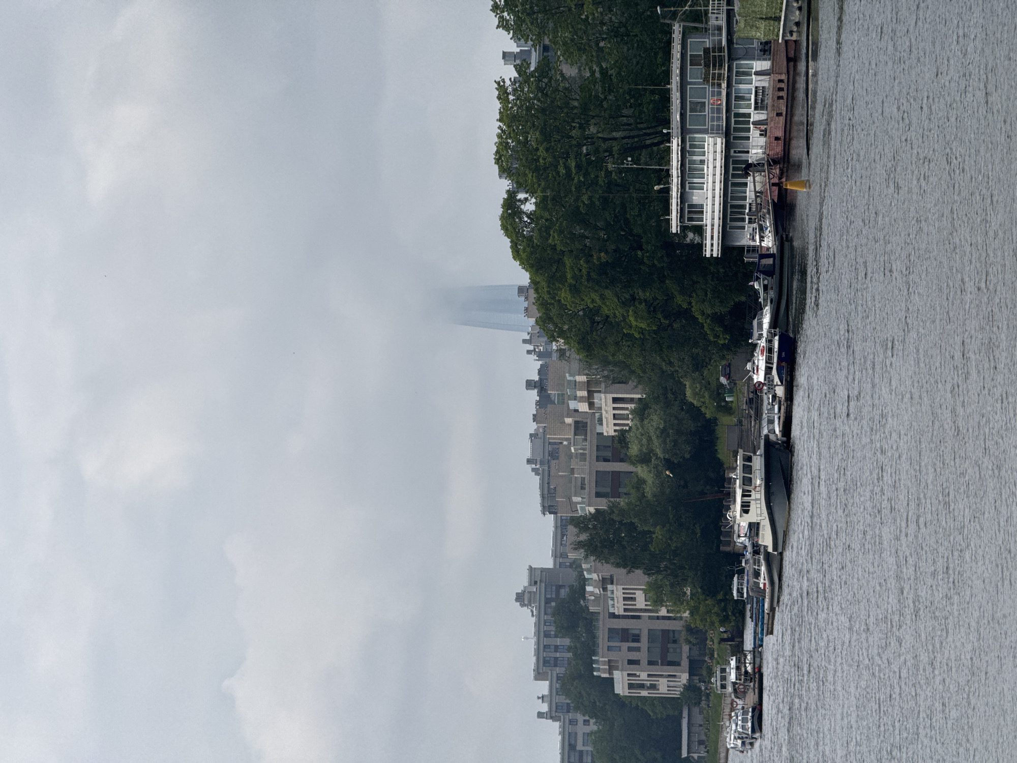 This is a photograph of a cloudy day on a body of water, likely a river or canal, in Saint Petersburg, Russia. In the foreground, the water is calm, with several boats moored along the shore. The boats are of various sizes and types, including what appear to be small passenger vessels and workboats.
Along the shore, there is a modern building with multiple levels and a long, narrow structure adjacent to it. Behind this building and extending further along the shore are several multi-story apartment buildings with a uniform, contemporary architectural style. Lush green trees are interspersed among the buildings, with some trees reaching considerable height and forming a dense canopy.
In the background, a very tall, slender skyscraper is visible, its upper portion obscured by heavy clouds. The overall atmosphere of the image is somewhat muted due to the overcast sky, creating a cool, diffused light. The scene suggests a residential or mixed-use area along the waterfront, with a mix of modern architecture and natural greenery. The presence of the distinctive skyscraper in the distance strongly suggests this location is Saint Petersburg. The skyscraper is the Lakhta Center.
