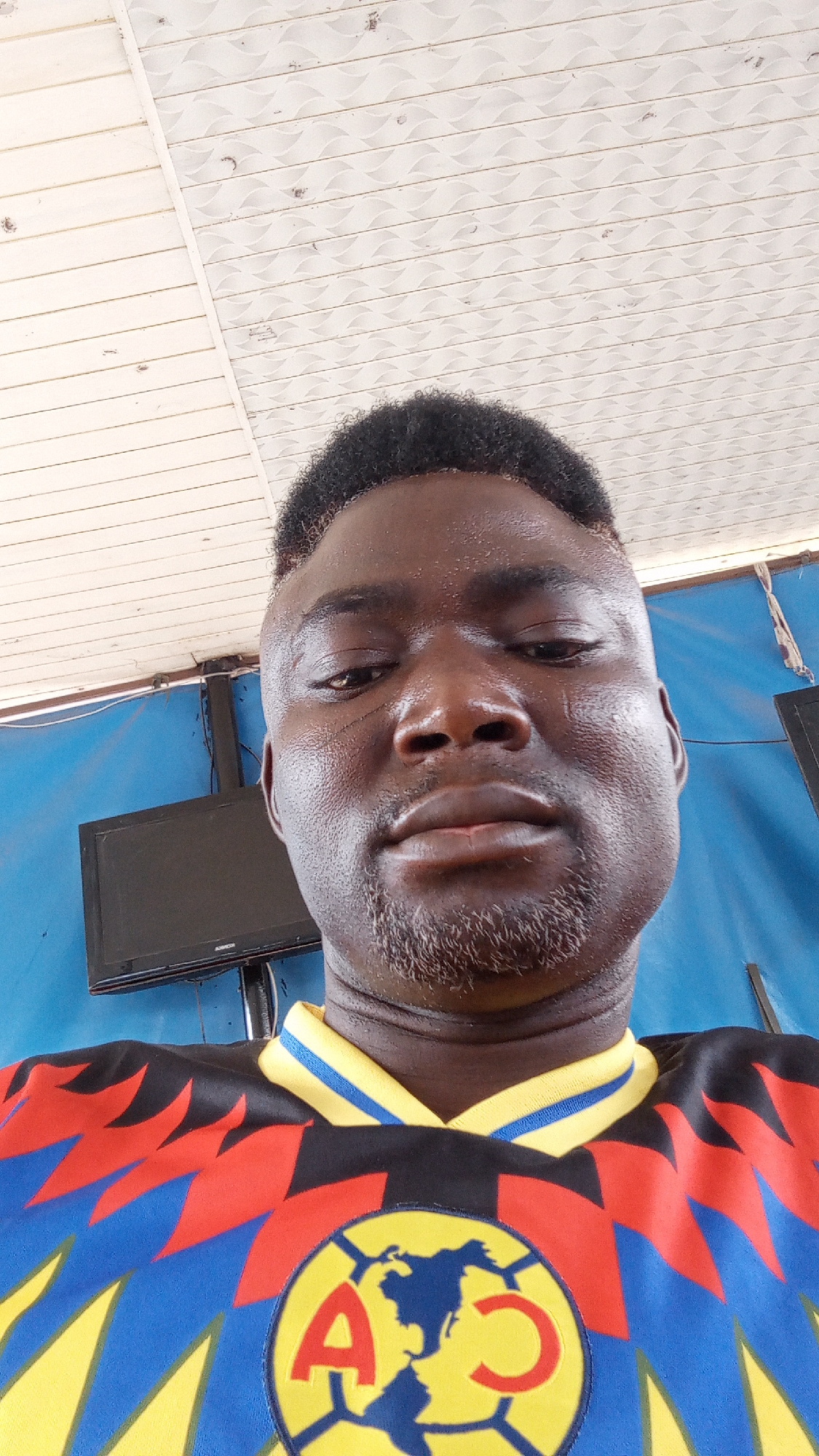 A selfie of a dark-skinned man. He has a short, dark haircut and a patchy beard. He is wearing a brightly colored Club America jersey with yellow, red, blue, and black geometric patterns. Behind him is a blue wall, and a small, dark-colored television is mounted on the wall. Above the man and behind the wall, there is a white ceiling with a textured pattern. The location is Sabon Tasha, Nigeria.