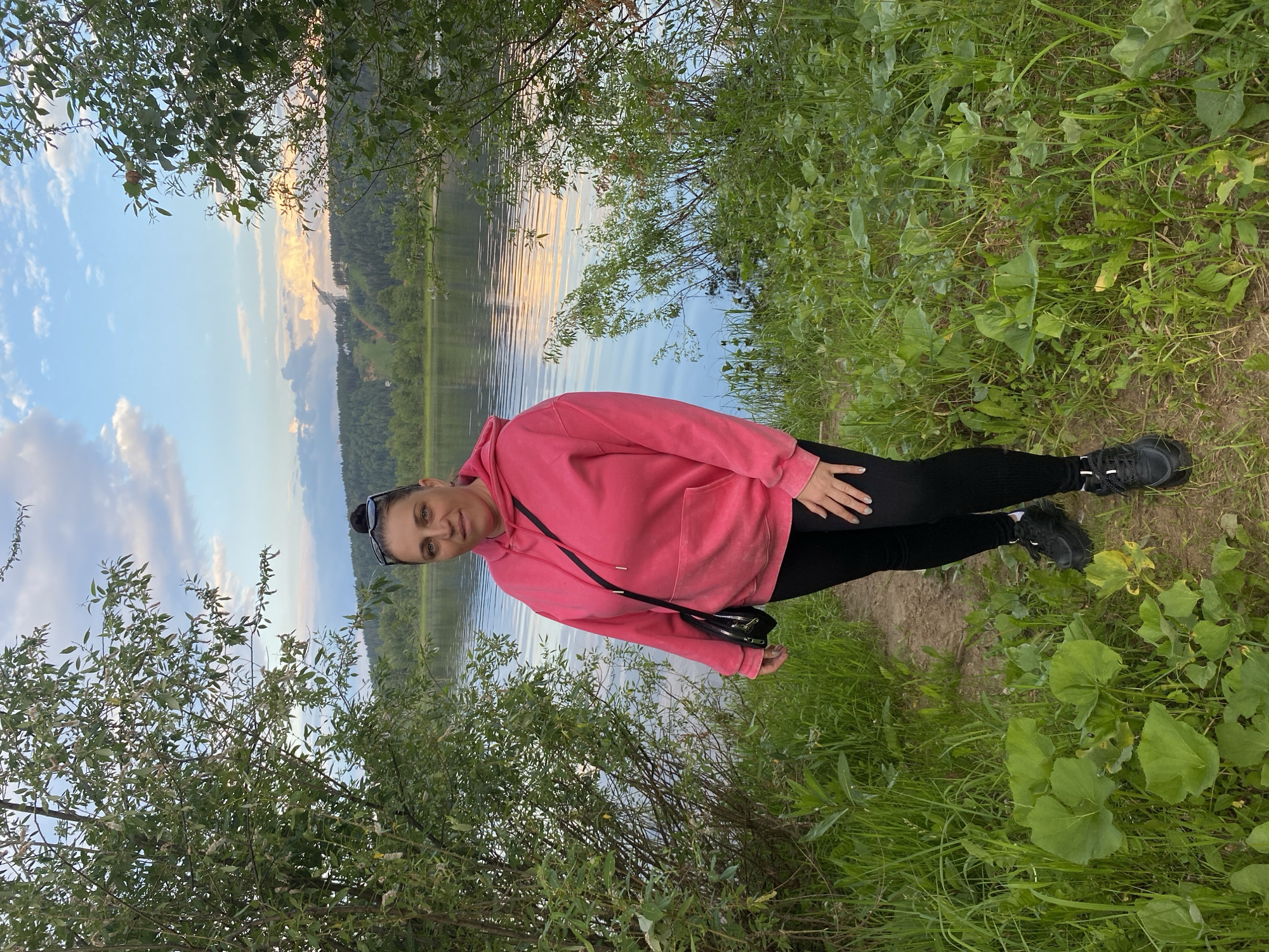 A woman is captured in a medium shot, posing in a pink hooded sweatshirt and black leggings. She is standing on a grassy bank beside a body of water, which appears to be a lake or river. The water reflects the sky, which is filled with clouds tinged with the warm hues of either sunrise or sunset. Trees and foliage line the bank and frame the water. In the distance, a forested hillside rises, suggesting a natural, possibly rural setting. The woman is looking towards the camera with a neutral expression. She has sunglasses perched on her head and a black purse slung across her body. The overall mood is peaceful and serene, evoking a sense of being outdoors in nature during a tranquil moment. The image orientation is portrait, with the subject appearing to be standing upright.