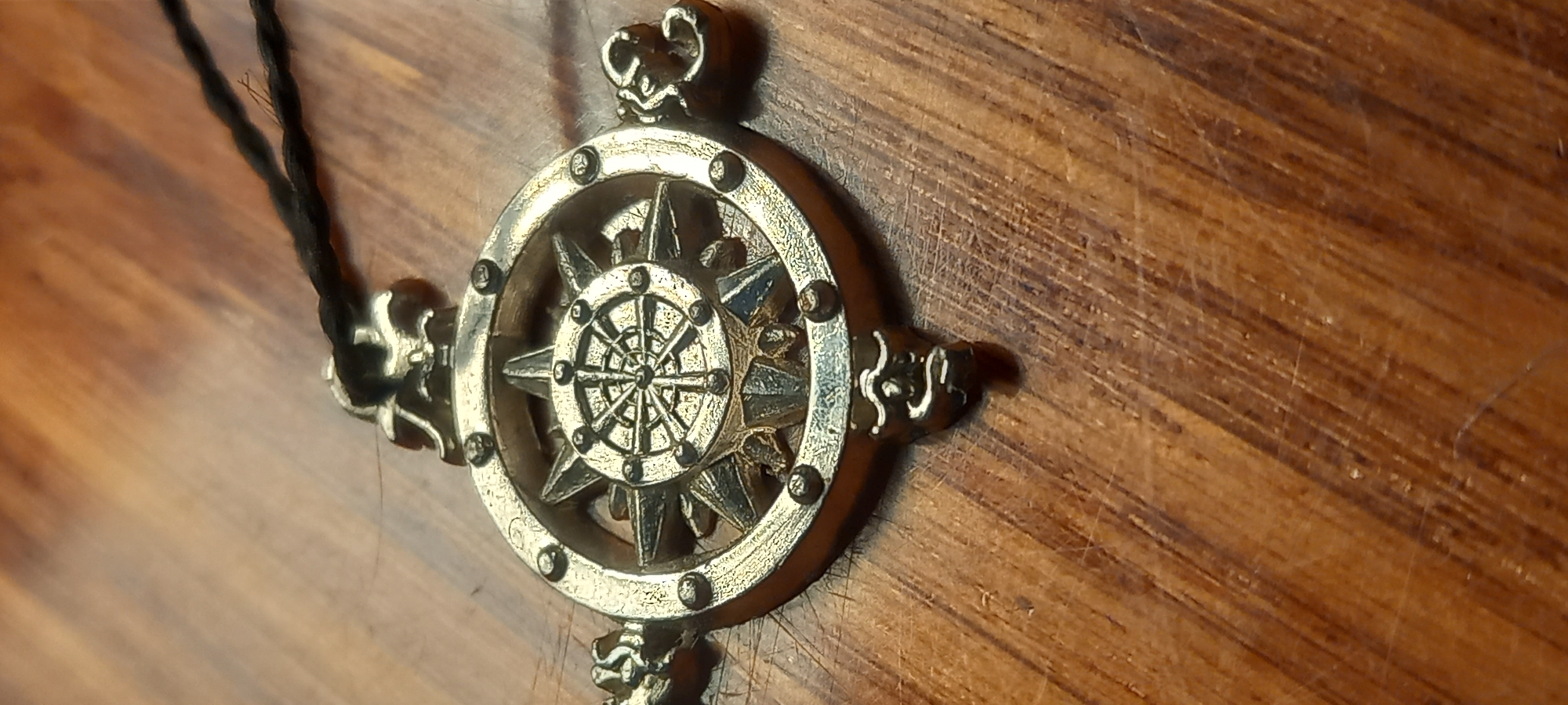 This is a close-up, eye-level, indoor shot of a weathered, ornate, metallic compass rose pendant lying on a wooden surface. The pendant is circular with an eight-pointed star design at its center, resembling a compass rose or a ship's wheel. The metal appears to be aged, with a textured, slightly tarnished finish, and it has a loop at the top for attachment to a cord. A black cord, twisted and frayed, is visible extending from the top of the pendant. The wooden surface is a medium-brown color with a distinct wood grain pattern, showing signs of wear and minor scratches. The lighting is somewhat dim, creating subtle shadows and highlighting the texture of the pendant and the wood. There are no people or other objects visible in the frame. The image's context does not provide information about its location in Kryvyi Rih, Ukraine, or any specific city.
