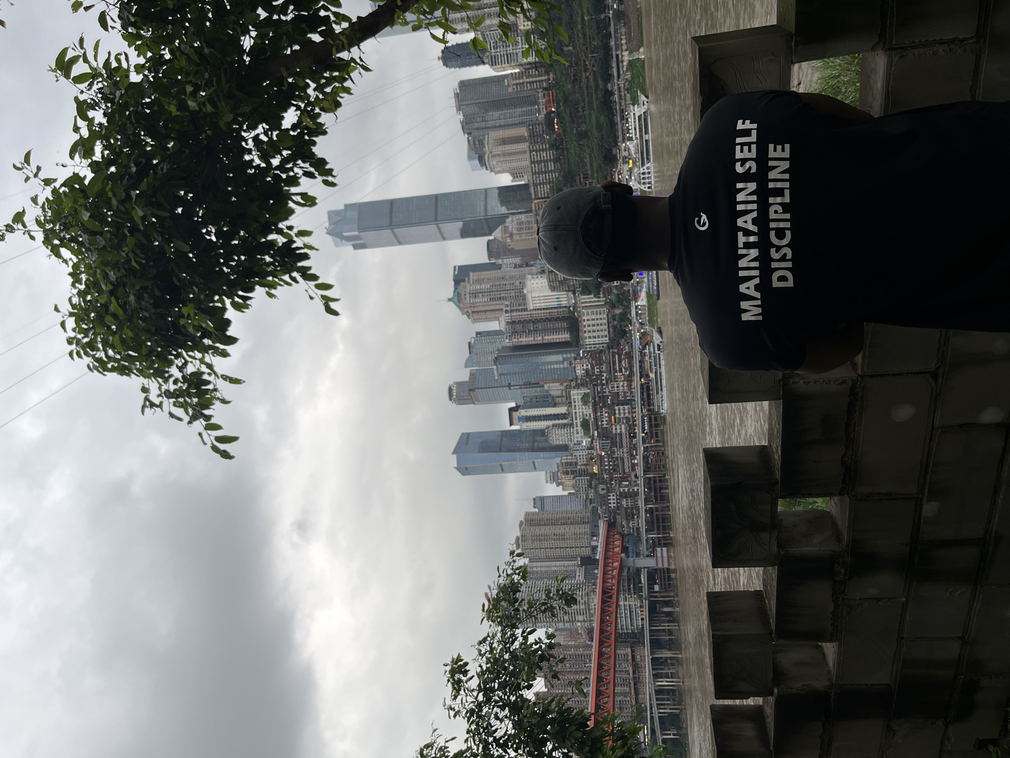 A person with a gray baseball cap is leaning on a brick wall overlooking a city skyline. The person is wearing a black t-shirt with white text reading "MAINTAIN SELF DISCIPLINE". The skyline includes a mix of skyscrapers and other buildings. An orange bridge is visible in the foreground. The sky is overcast. The location is the Yuzhong District of Chongqing, China. Green foliage from trees is present on either side of the frame.