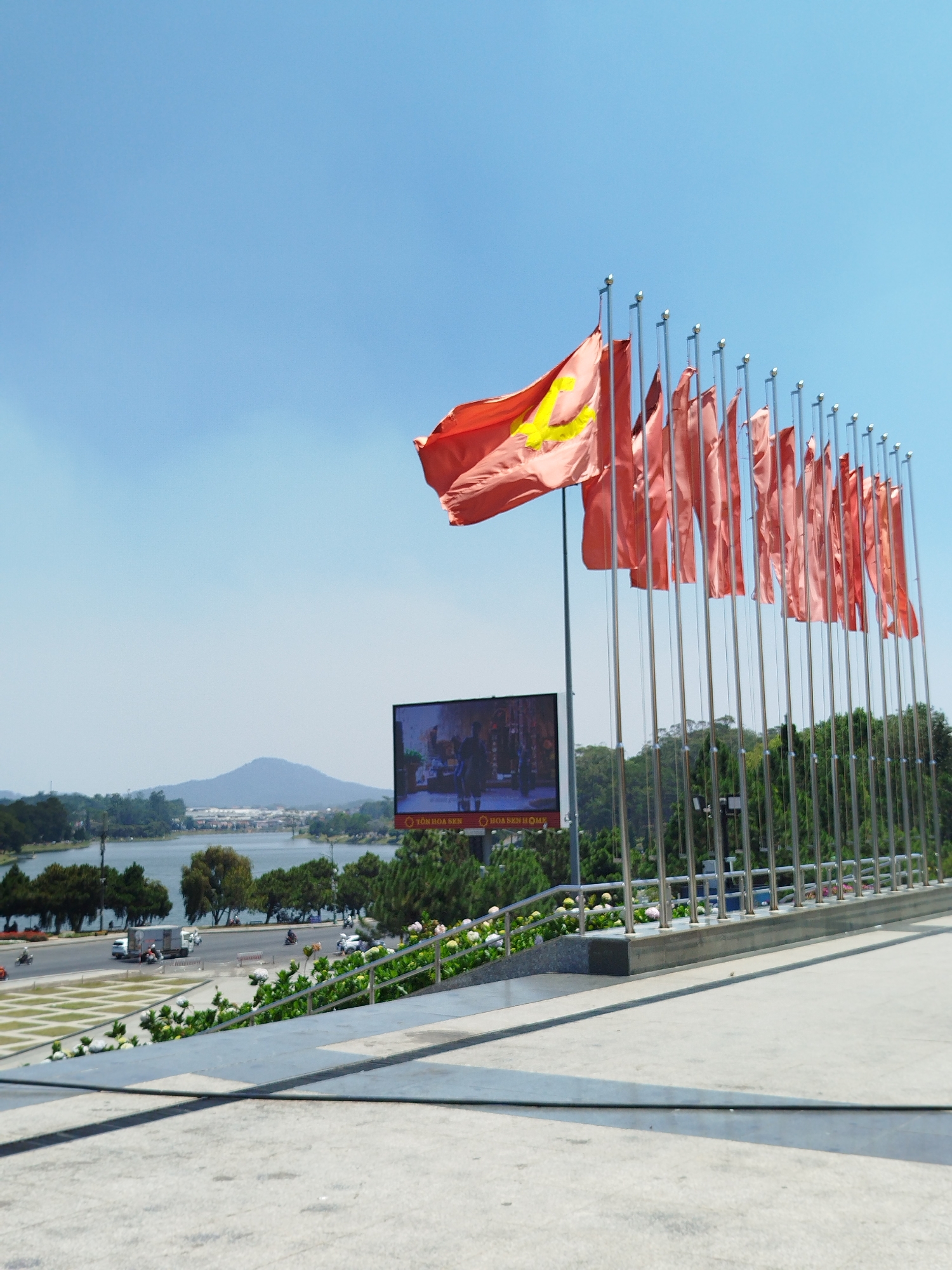 A ground-level shot in Da Lat, Vietnam, showcasing a row of approximately 20 red flags affixed to metal poles, fluttering in the wind. The first flag displays a gold star. Behind the flags is a large video screen showing figures. Further back is a body of water, with trees and a low mountain range in the background, under a clear blue sky. A road with vehicles is visible to the left of the screen. The foreground shows a concrete surface.