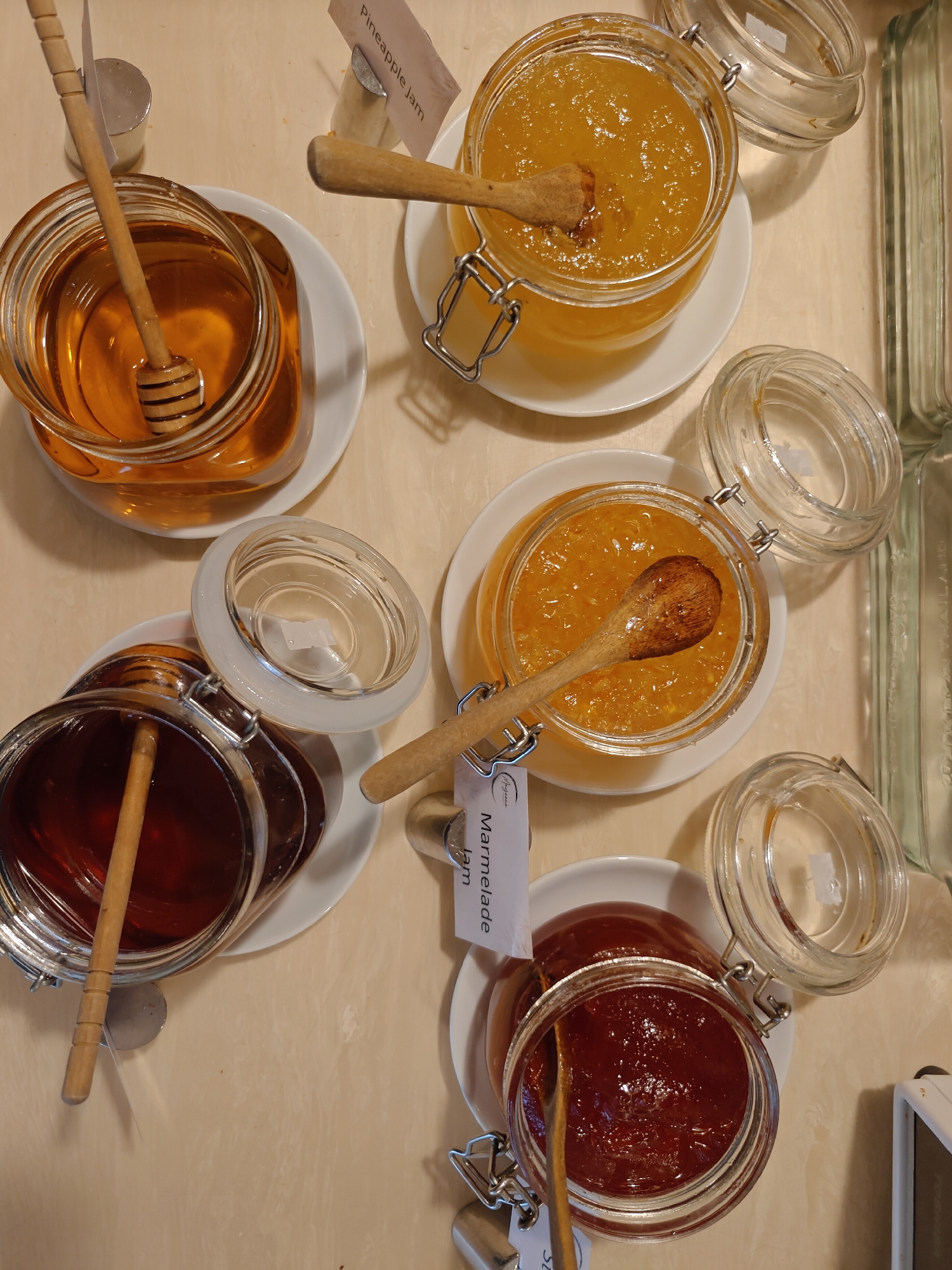 An overhead, close-up shot depicts an assortment of jams and honey in glass jars with wooden stirrers. The scene is set in Pattaya City, Thailand, on a light-colored, textured surface that appears to be a table.
In the upper portion of the image, a jar filled with golden honey is positioned on a white saucer. A wooden honey dipper rests within the jar. To its right, another glass jar contains a bright yellow jam, likely pineapple, also on a white saucer with a wooden stirrer. A label indicating "Pineapple Jam" is visible.
Below the honey and pineapple jam, a dark amber-colored jam or preserve is presented in a glass jar, resting on a white saucer. A wooden stirrer is placed inside this jar, and a label that reads "Marmalade Jam" is attached to the jar's neck.
To the right of the marmalade jam, a jar with a dark red jam or preserve is visible, its lid open and a wooden stirrer inserted. This jar is also on a white saucer.
Several empty glass jar lids are scattered around the jars. The overall impression is one of a breakfast or buffet setting offering a selection of sweet spreads. The lighting is soft and diffused, highlighting the textures and colors of the jams and honey.