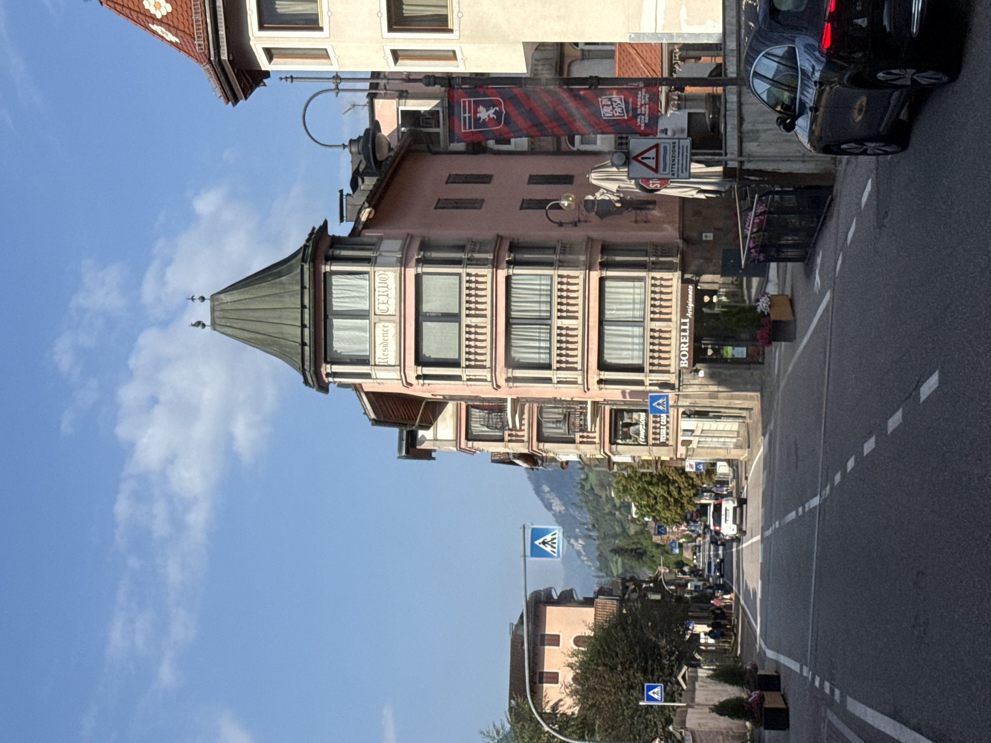 In Moena, Italy, on a sunny day with a partly cloudy sky, a street scene unfolds with buildings lining both sides of the road. The architecture features a blend of traditional and modern styles, with pastel-colored facades, balconies, and ornate window frames. A prominent building on the right displays a banner with the words "VAL DI FASSA."
On the street, traffic moves along a paved road marked with white lines. Several cars are visible, including a black SUV in the foreground and a white car further down the road. Pedestrians are present, walking along the sidewalks and crossing the street at designated crosswalks, indicated by blue and white pedestrian crossing signs. Lush green trees and potted plants add to the urban landscape. The backdrop features rolling hills and mountains, suggesting a scenic, mountainous region.