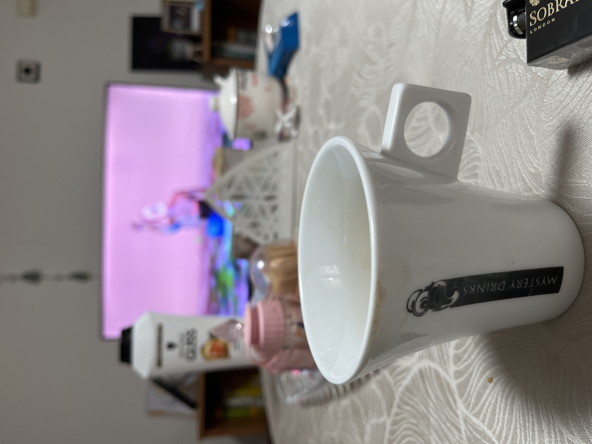 A white ceramic mug, angled to the right, is the primary subject, bearing a black label that reads "MYSTERY DRINKS." The mug has a square-shaped handle with a circular opening. Behind the mug, a table covered with a patterned white tablecloth hosts various blurred objects including a small pink bottle, a white carton, and a decorative white container. In the background, a television screen displays a brightly lit, abstract image. A wooden shelf unit is visible partially behind the television. A "SOBRANIE LONDON" box sits near the top right corner of the frame. The location and date are not determinable from the image. The presence of Bulgarian text is not determinable from the image.