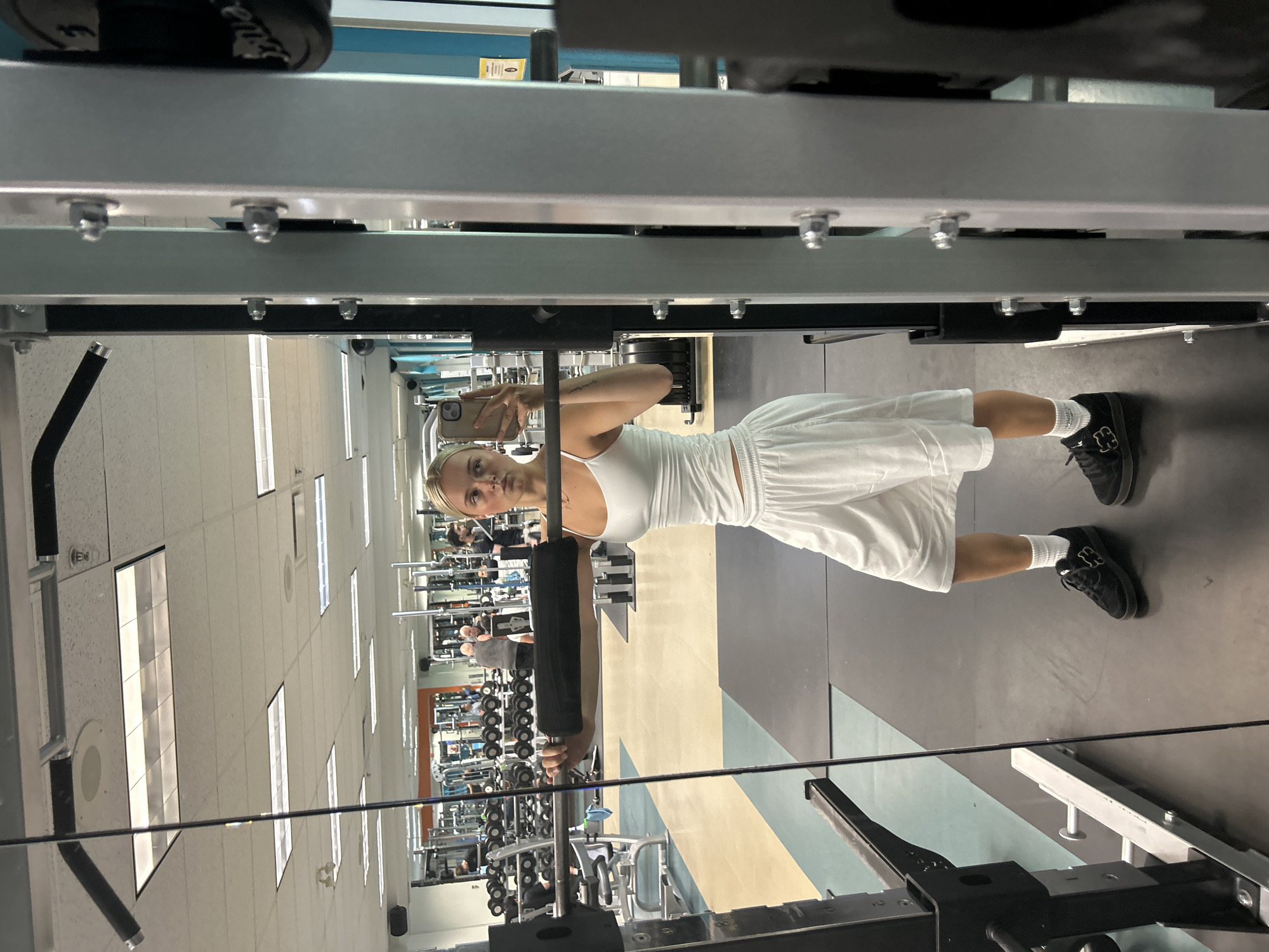 The image depicts a young woman in a gym, reflected in a mirror. She is wearing a white tank top, white shorts, white socks, and black athletic shoes. She is holding a barbell with a padded attachment across her upper back, suggesting she is preparing for or in the middle of a weightlifting exercise. The gym environment is visible in the background, with weight racks, other gym equipment, and several other individuals working out. The lighting appears to be typical for an indoor fitness facility, with fluorescent ceiling lights. The image does not contain any identifiable landmarks or contextual clues that would place it specifically in Whistler, Canada.