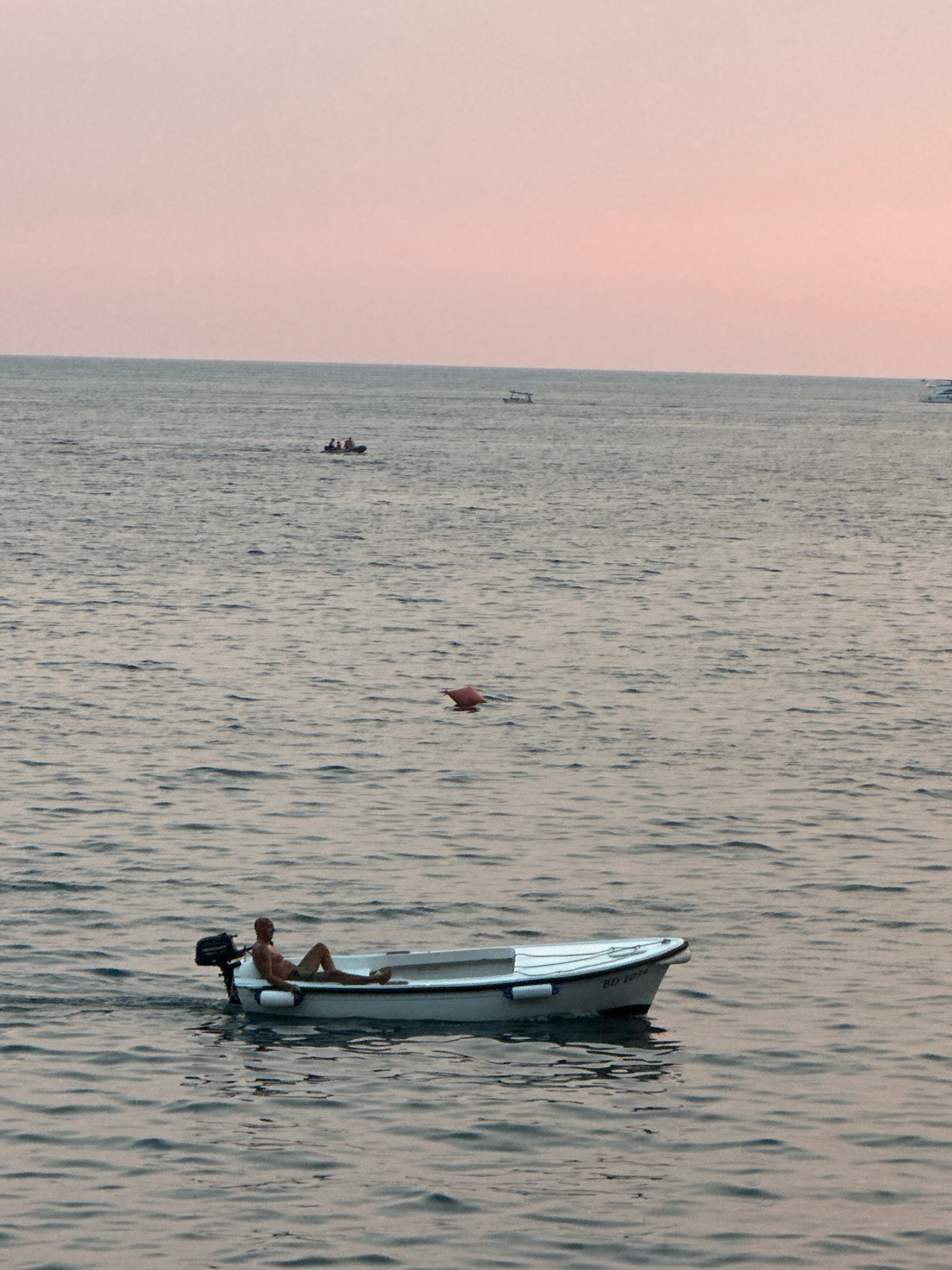 A person is reclining in a small white boat with an outboard motor in the Adriatic Sea off the coast of Budva, Montenegro. The boat has the registration number "BD 1034" visible on its hull. The water is calm with gentle ripples. In the distance, other boats are visible, including a dinghy with several people in it and a larger vessel. A pink buoy floats in the water to the right of the boat with the person. The sky is a soft pink, indicating either sunrise or sunset.
