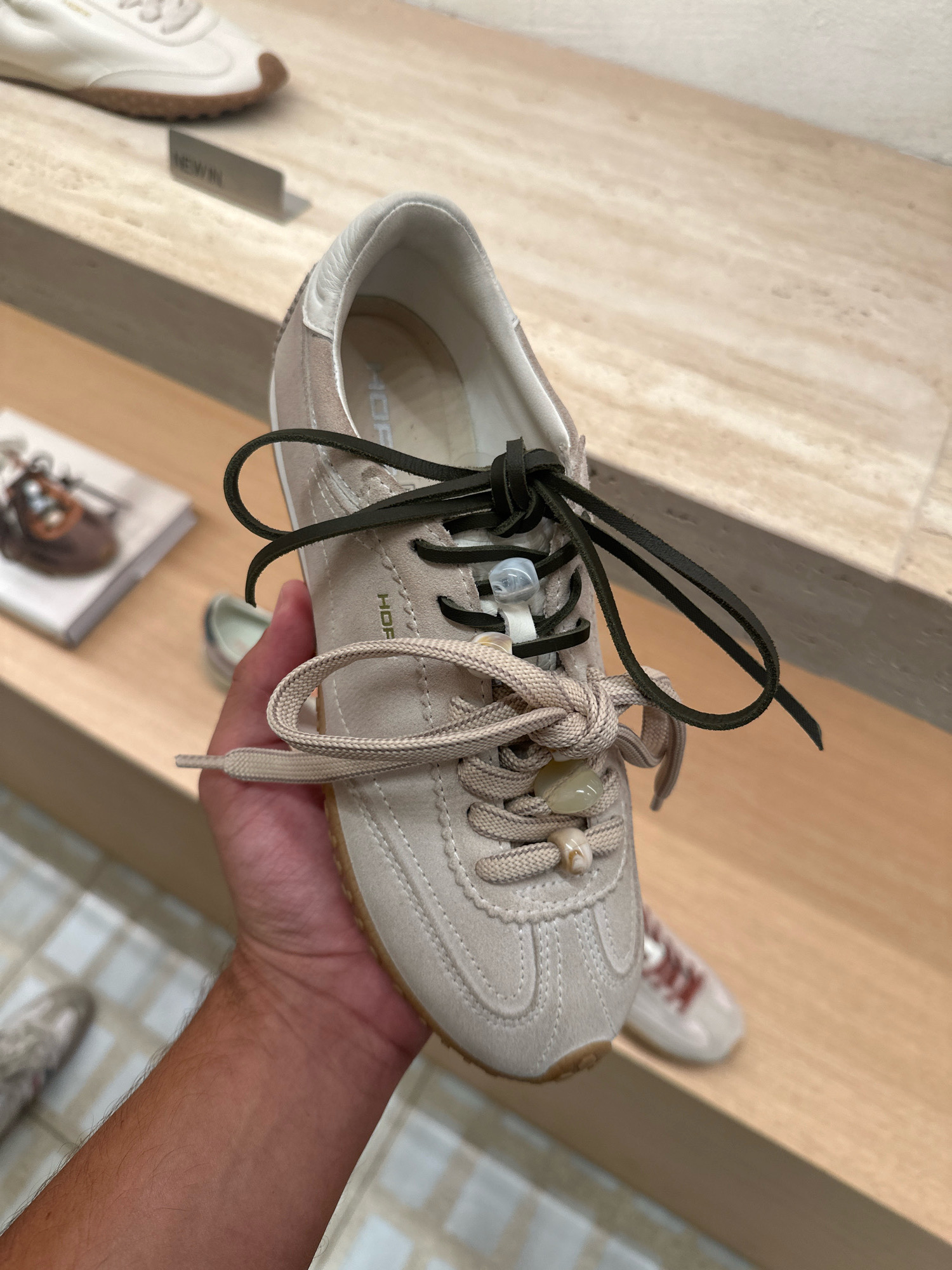 A person's hand holds a close-up view of a beige low-top sneaker with two sets of laces. One set is a dark green leather lace threaded through the eyelets and tied with a small clear plastic toggle. The other set is a beige braided lace, also threaded through the eyelets and tied with a knot, adorned with two light-colored natural stone beads. The sneaker has a white leather trim around the ankle and a tan sole. In the blurred background, other shoes are visible on wooden shelves, including a white sneaker with a brown sole and another beige sneaker with red laces. A small sign that reads "NEW IN" is also visible in the background. The scene appears to be inside a retail store.