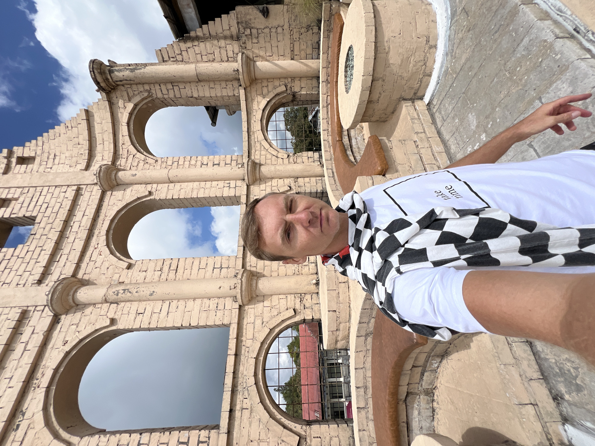 A man takes a selfie in front of a sun-baked brick structure with arched openings, possibly a ruin or themed building, in Kintamani, Indonesia. He is wearing a white t-shirt with the text "take time" and a black and white checkered scarf draped over his shoulders. The structure features stone columns and semicircular seating around a central fire pit. In the background, through the arched openings, glimpses of a red-roofed building and trees can be seen, as well as a blue sky with scattered clouds. The overall scene suggests an outdoor location with a rustic, possibly historical, ambiance.