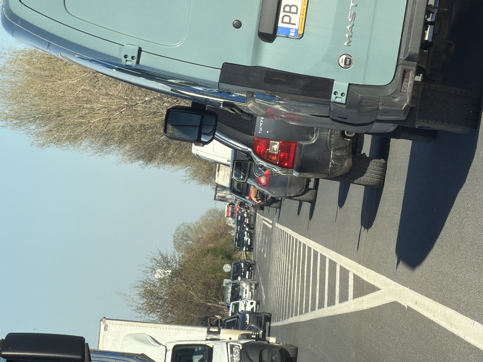 The image shows a traffic jam on a road, with several vehicles lined up in a row. The foreground features a light blue van with a Bulgarian license plate "PB" and "MASTER" written on the back. Adjacent to the van is a dark gray "SPORT" SUV. In the distance, there is a mix of cars and trucks, suggesting possible congestion on a highway or a main road. The road markings include a series of white diagonal lines marking a crosswalk. Trees line the side of the road, and the sky is clear blue, indicating it's daytime. Without additional landmarks, I cannot definitively confirm the location within Plovdiv, Bulgaria.