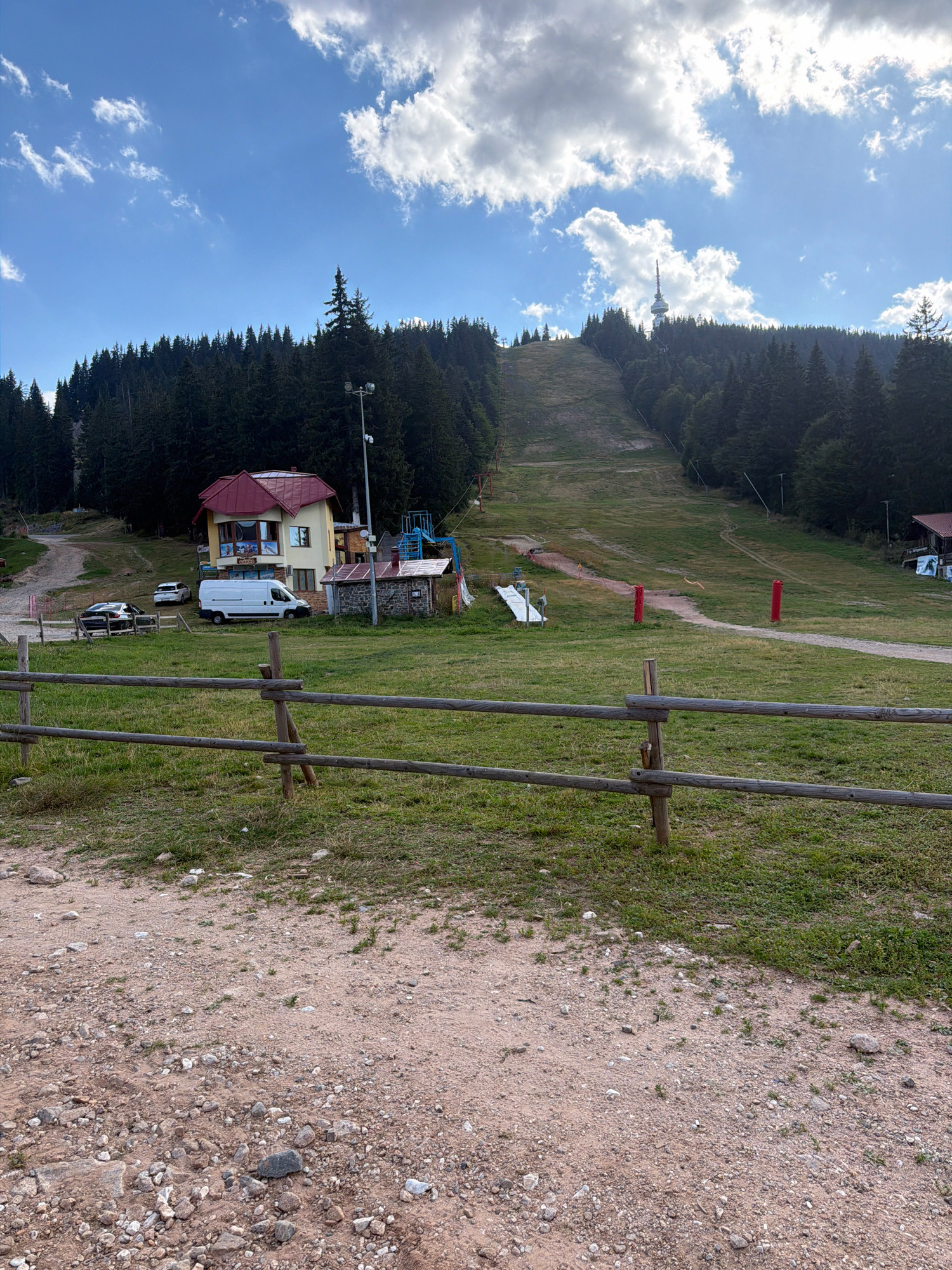 This image depicts a view of a ski slope in the city of Smolyan, Bulgaria, during a clear day with scattered clouds. In the foreground, a rustic wooden fence runs horizontally across the frame, separating a gravelly path from a grassy field. Behind the fence, a yellow building with a red roof is visible, accompanied by a smaller stone structure. Several vehicles, including a white van and two cars, are parked near the buildings. The ski slope itself is a wide, grassy expanse stretching uphill, bordered by dense evergreen trees. A tall communication tower is observable on the horizon, atop a tree-covered hill. The overall scene suggests a recreational area in a mountainous region.