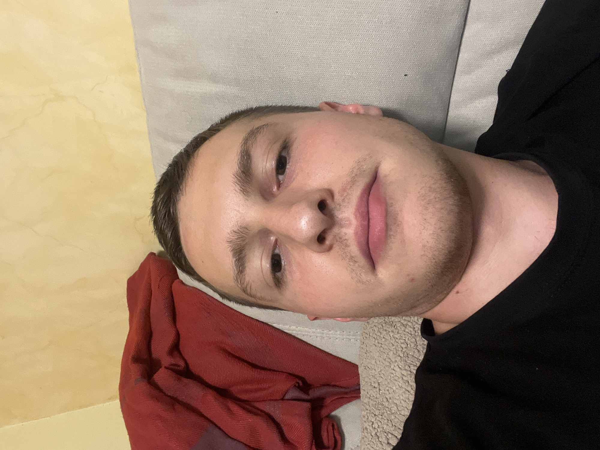 A fair-skinned young man with short, dark hair and stubble is lying down, his head resting on a beige textured surface, possibly a sofa or cushion. He is wearing a black top. To the left of the frame, a red crumpled cloth is visible. The background consists of a yellowish wall with apparent surface variations. The location cannot be ascertained as Burgas, Bulgaria, based on the visual information provided.