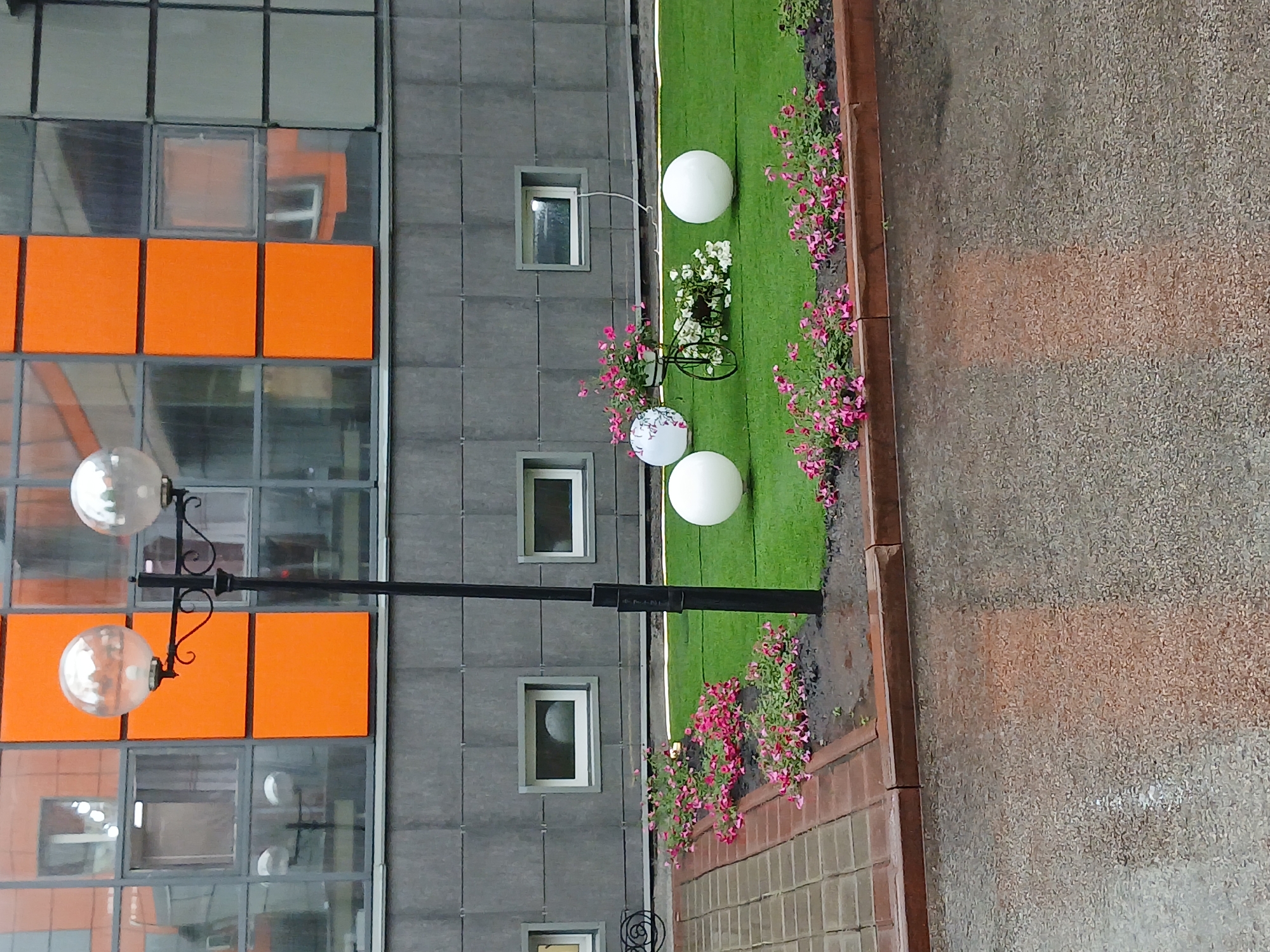 The image shows an outdoor decorative scene in front of a modern building with an orange and grey facade. A black lamppost with two spherical glass light fixtures stands in the center. The ground is covered with artificial green turf, bordered by red brick pavers and a planter with pink flowers. There are three large white spherical decorations placed on the turf, and a small decorative bicycle with a pot of flowers. The building has multiple windows, some of which reflect the surroundings. The pavement in the foreground is wet, indicating recent rain. This scene is located in the city of Kotino, Russia.