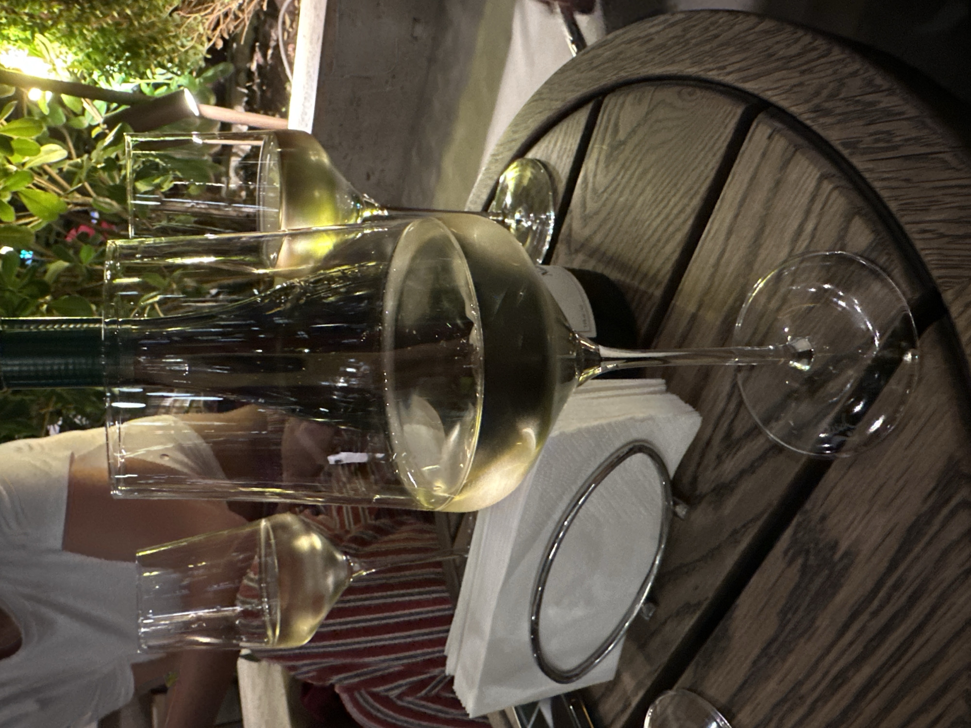 A nighttime photograph depicts a restaurant scene in Tbilisi, Georgia. A wooden table takes up the majority of the frame, upon which rest a half-full bottle of white wine, two wine glasses filled with white wine, and a stack of white napkins in a silver holder. A person wearing a white t-shirt is partially visible in the left foreground. In the background, green plants are illuminated by a light.