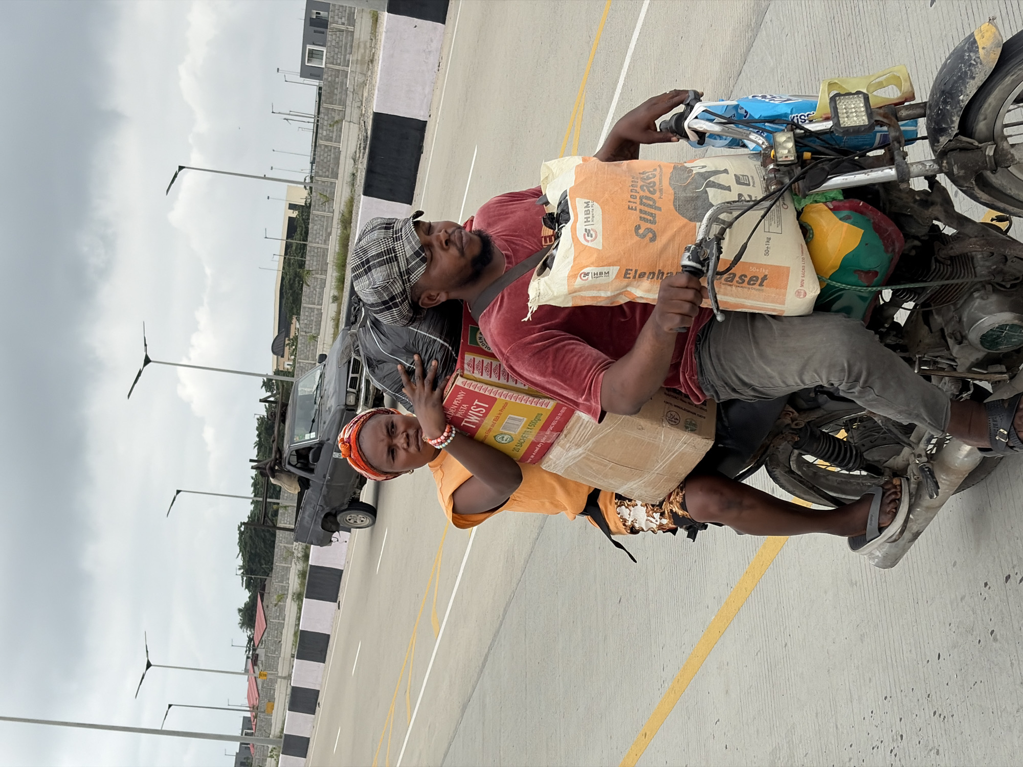 An overcast day in Igboefon, Nigeria, shows a man and woman riding a motorcycle laden with cargo on a paved road. The man, wearing a red t-shirt and grey pants, drives the motorcycle, his hands on the handlebars. He wears a checkered flat cap. A large orange bag with an elephant logo, labeled "Elephant Supa," is strapped to the front of the motorcycle. The woman, in an orange sleeveless top and a patterned skirt, sits behind him, holding onto a large cardboard box. Another cardboard box is strapped to the back of the motorcycle. The road is marked with yellow and white lines. In the background, a black pickup truck drives ahead, and streetlights and some buildings are visible.