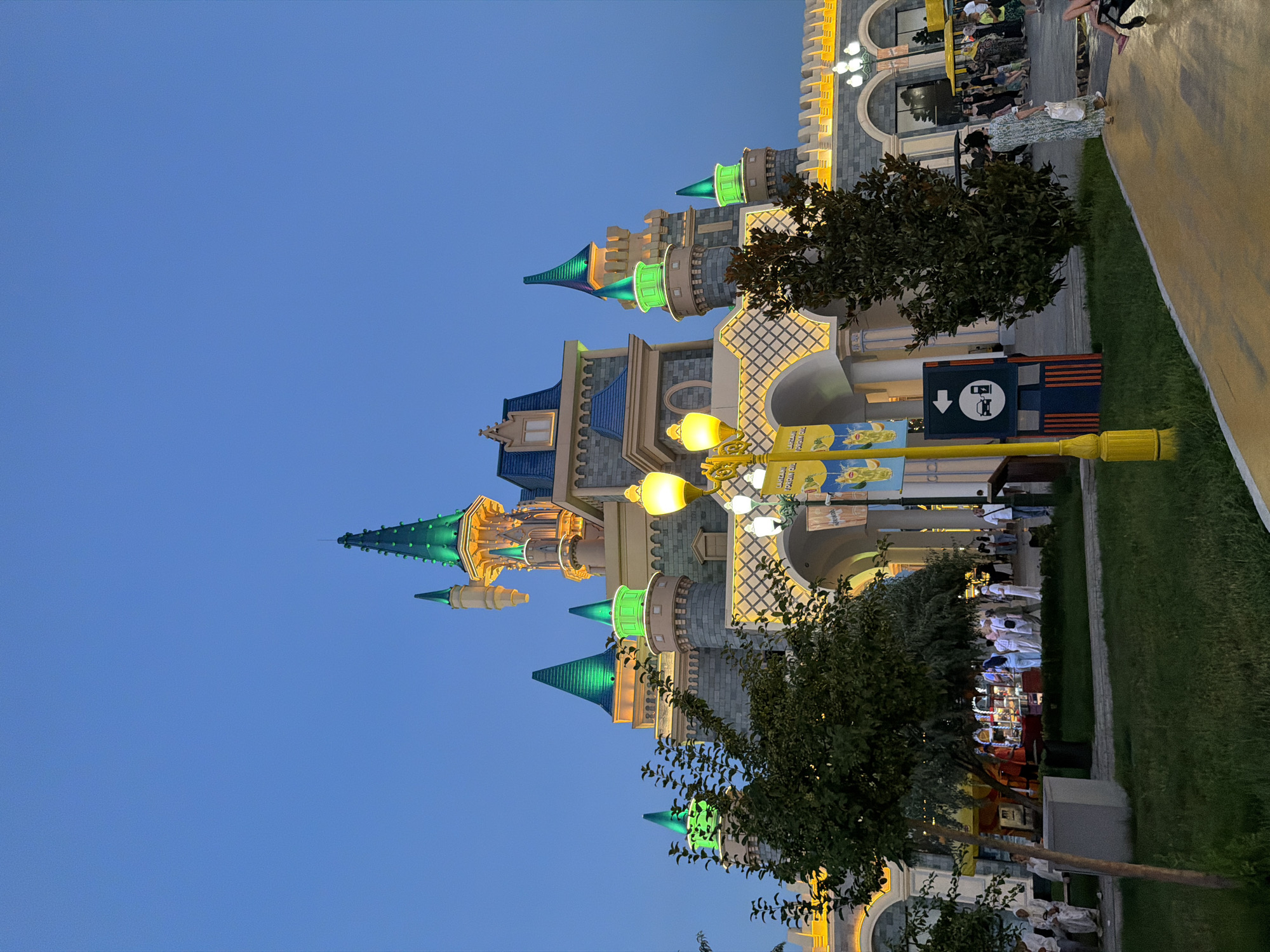 This is a vertical, full-shot photograph of a whimsical, fairy-tale-style castle at dusk, likely in an amusement park or entertainment complex. The castle features multiple conical turrets, some illuminated with vibrant green lights, others with warm yellow light. The architecture is a blend of stone-like materials and decorative elements, reminiscent of a fantasy kingdom.
In the foreground and midground, several people are visible, scattered along a paved pathway and grassy areas. They appear to be strolling, with some gathered in small groups. The activity suggests a leisurely evening at a public attraction. The lighting indicates it is twilight, with artificial lights illuminating the castle and pathway, creating a festive and magical atmosphere. The sky is a clear, deep blue, typical of an evening with fair weather.
To the right of the castle, a yellow lamppost with decorative embellishments stands prominently. Attached to it are signs, one of which is a blue square with white graphics indicating a bus stop or transport hub, with an arrow pointing downwards. Another sign above it features yellow and blue stripes with some illegible text. Further to the right, a wide, light yellow pathway curves out of frame. A lush green lawn borders the pathway, with trees providing shade and visual interest.
The scene evokes a sense of wonder and enjoyment, common in places designed for entertainment and recreation. The overall impression is one of a charming evening outing. The location context suggests this could be a landmark or attraction in Tashkent, Uzbekistan.