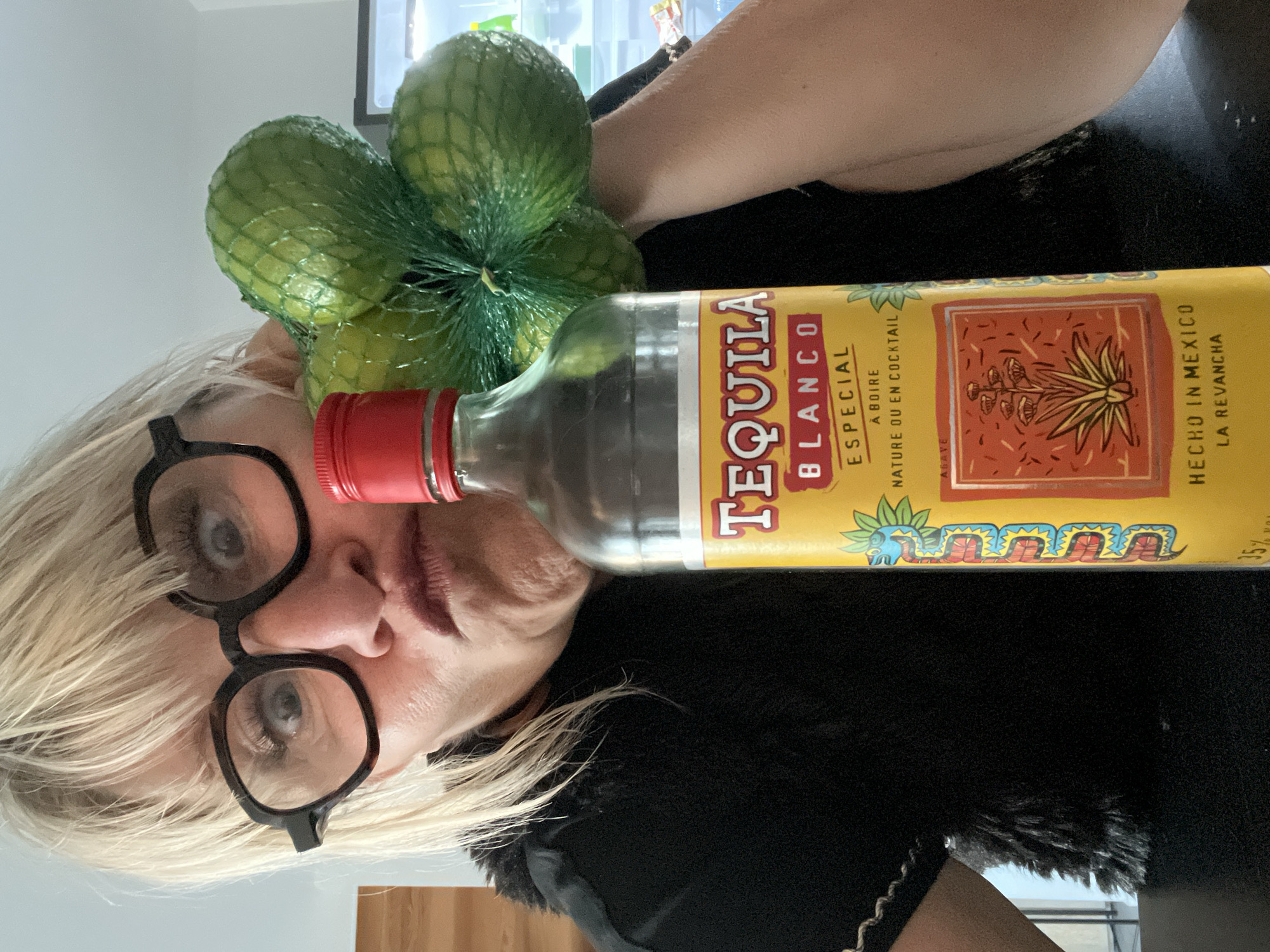 The image, correctly oriented, presents a close-up of a blonde-haired person with black-framed glasses, partially obscured by a bottle of tequila and a net bag of limes. The person's eyes are visible above the glasses, looking slightly upwards, and their mouth has a neutral, slightly pouted expression. They are wearing dark, possibly furry, clothing.
A clear bottle of "TEQUILA BLANCO ESPECIAL" with a red cap is prominently featured in the foreground. Its yellow label displays "A BOIRE NATURE OU EN COCKTAIL," "AGAVE," "HECHO IN MEXICO," "LA REVANCHA," and "35%." The label also includes illustrations of an agave plant and a stylized serpent. Behind the tequila bottle and near the person's head, a green mesh bag contains several vibrant green limes.
The scene is set indoors, likely a casual home environment in Chambœuf, France, indicated by light-colored walls and an out-of-focus background. The arrangement of the tequila bottle and limes suggests the person is either preparing or presenting ingredients for a cocktail, possibly a margarita. The lighting indicates an indoor setting, without specific cues for the time of day or weather. The image captures a moment of poised presentation of these items.