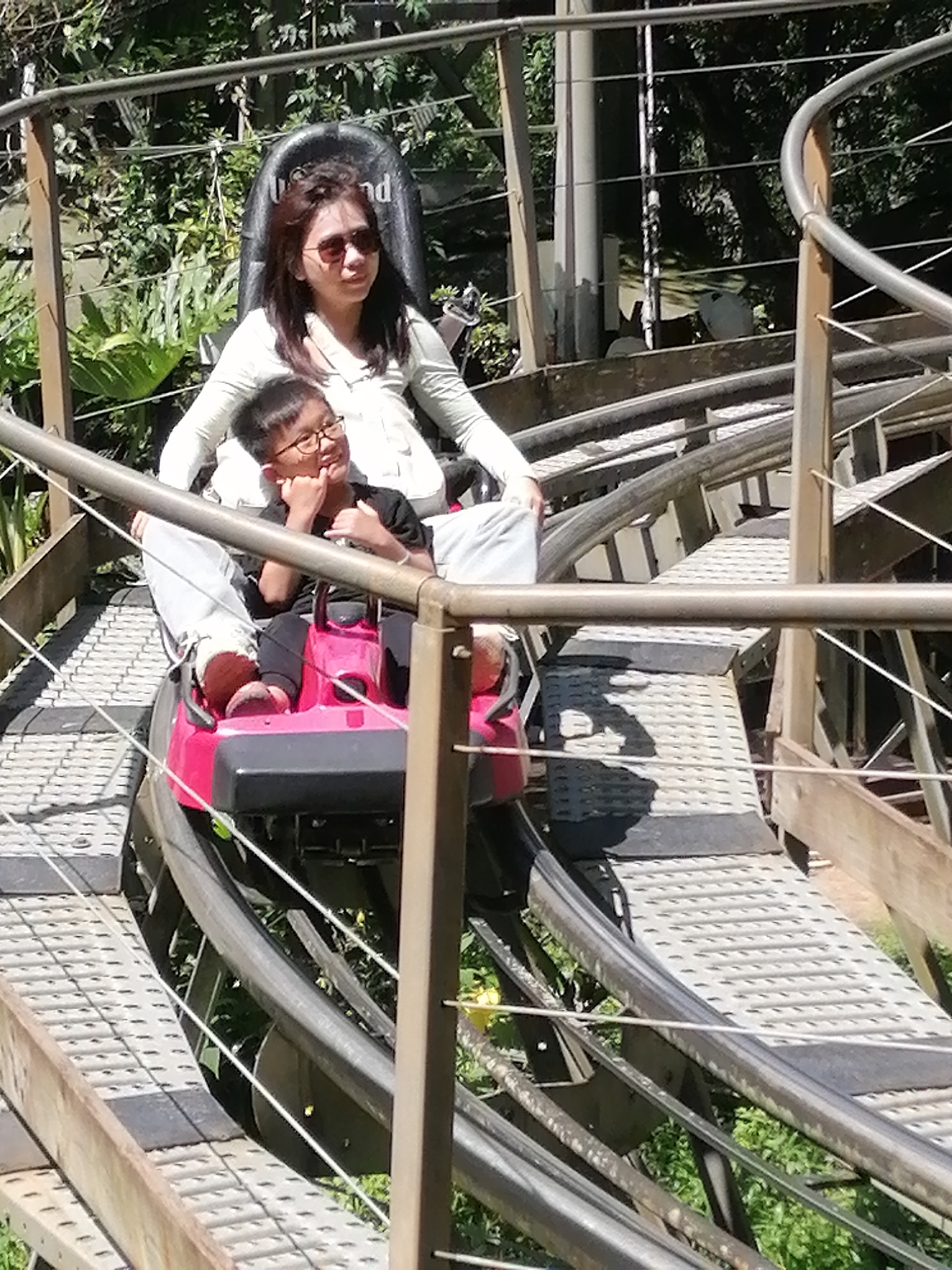 This media file shows two people on a scenic rollercoaster ride. The subjects are a woman and a young boy. The woman, in the front seat, is wearing sunglasses and a light-colored long-sleeved shirt. She appears to be looking towards the right. The boy, seated behind her, is wearing a black t-shirt and glasses. He is looking towards the left, with his hand touching his chin, and appears to be smiling or amused. They are both seated in a bright pink rollercoaster car with black seats. The rollercoaster track is made of metal rails and has a grated metal walkway. The surrounding environment is lush and green, with trees and foliage visible in the background, suggesting an outdoor setting with natural beauty. The lighting suggests it is daytime, likely sunny given the shadows. The image appears to be taken in Da Lat, Vietnam, known for its scenic attractions. Visible text on the seat headrest reads "nd". The overall mood seems happy and adventurous.