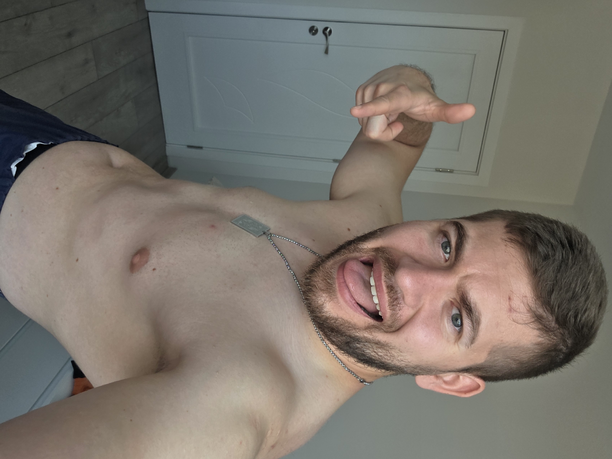 A shirtless man with a beard and blue eyes is pointing his finger at the viewer. He is wearing a dog tag necklace and appears to be sticking his tongue out. The background includes a white door and light grey flooring. The location is indeterminable, there is nothing in the image to suggest Yagodovo, Bulgaria.