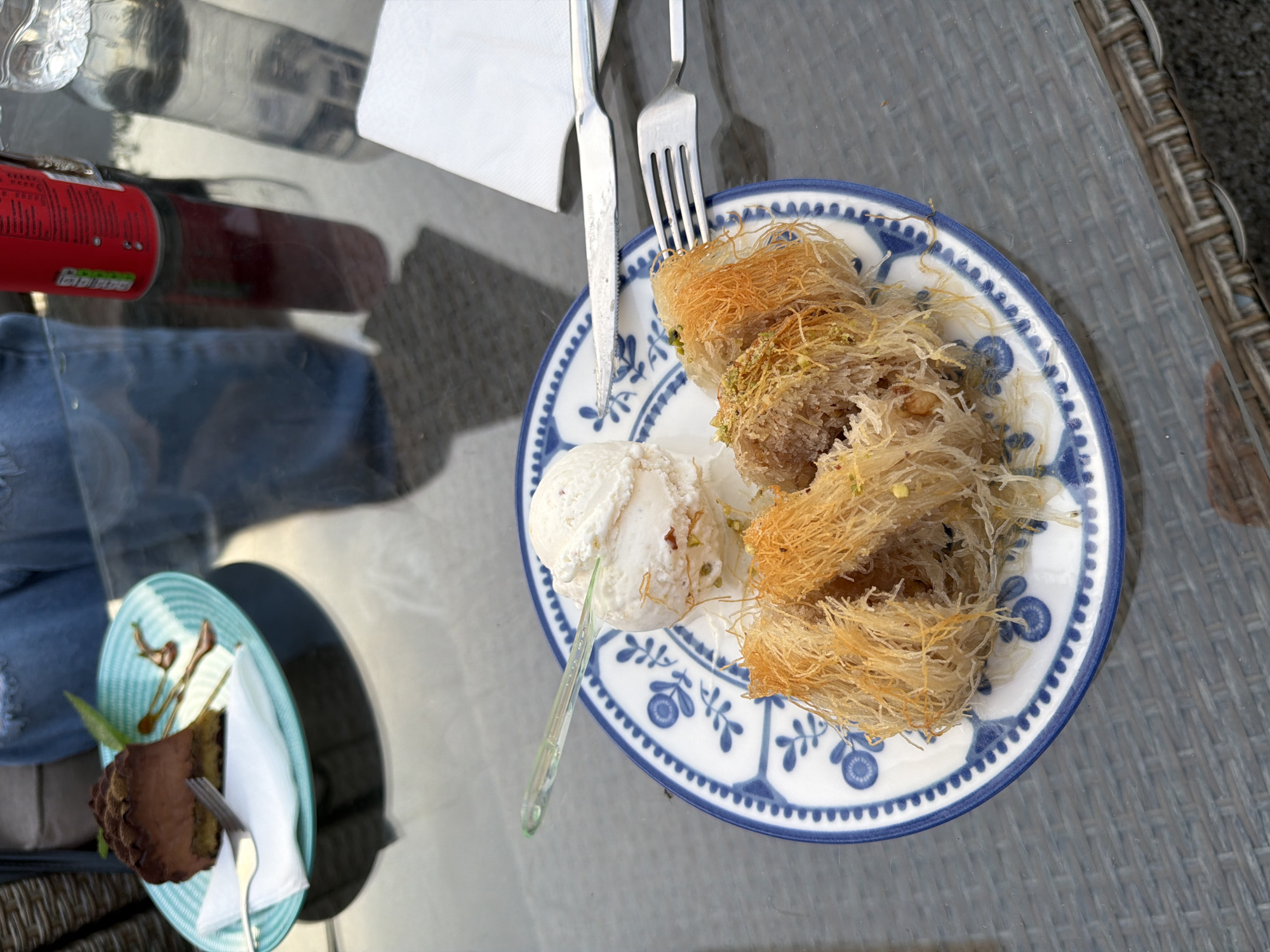 The image depicts a meal setting, likely outdoors, on a glass-topped table with a woven rattan-like frame along the edge. On the table, there's a plate with a blue and white patterned design. The plate holds several pastries resembling shredded phyllo dough nests, and a scoop of ice cream. Utensils (a knife and fork) rest on the plate next to a folded white napkin. In the background, there's a red and white can, a clear bottle partially filled with liquid, and a glimpse of a person wearing denim. Further left, there's another plate with a slice of chocolate cake accompanied by a white napkin and fork.
The location is likely Nesebar, Bulgaria, as the presence of Kataifi pastry, a traditional dessert in Balkan cuisine, points to this region.