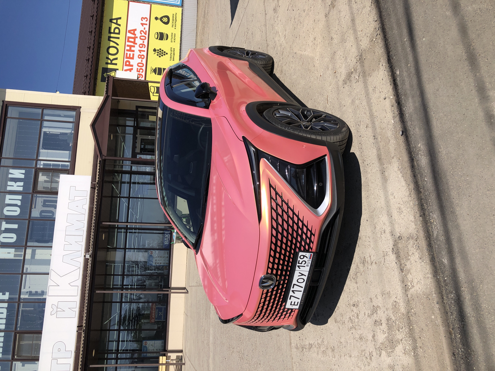 The image captures a pink Changan UNI-K iDD plug-in hybrid car parked on a paved surface in what seems to be an urban environment, specifically in the city of Chaikovsky, Russia. The car is facing the viewer, with its license plate "E717OY 159" clearly visible.
Behind the car, a building with large windows and a sign reading "ТР Й КЛИМАТ" is visible. Above this is a smaller sign that reads "КОЛБА АРЕНДА 950-819-02-13" followed by icons representing goods for winemaking and various containers, as well as "РАСХОДНИКИ И ИНГРЕДИЕНТЫ" which translates to " расходники и ingredients."
The sky is blue and clear, suggesting the photo was taken on a sunny day. There are shadows on the pavement, indicating the position of the sun. No people are visible in the image. The environment suggests a commercial or retail area.