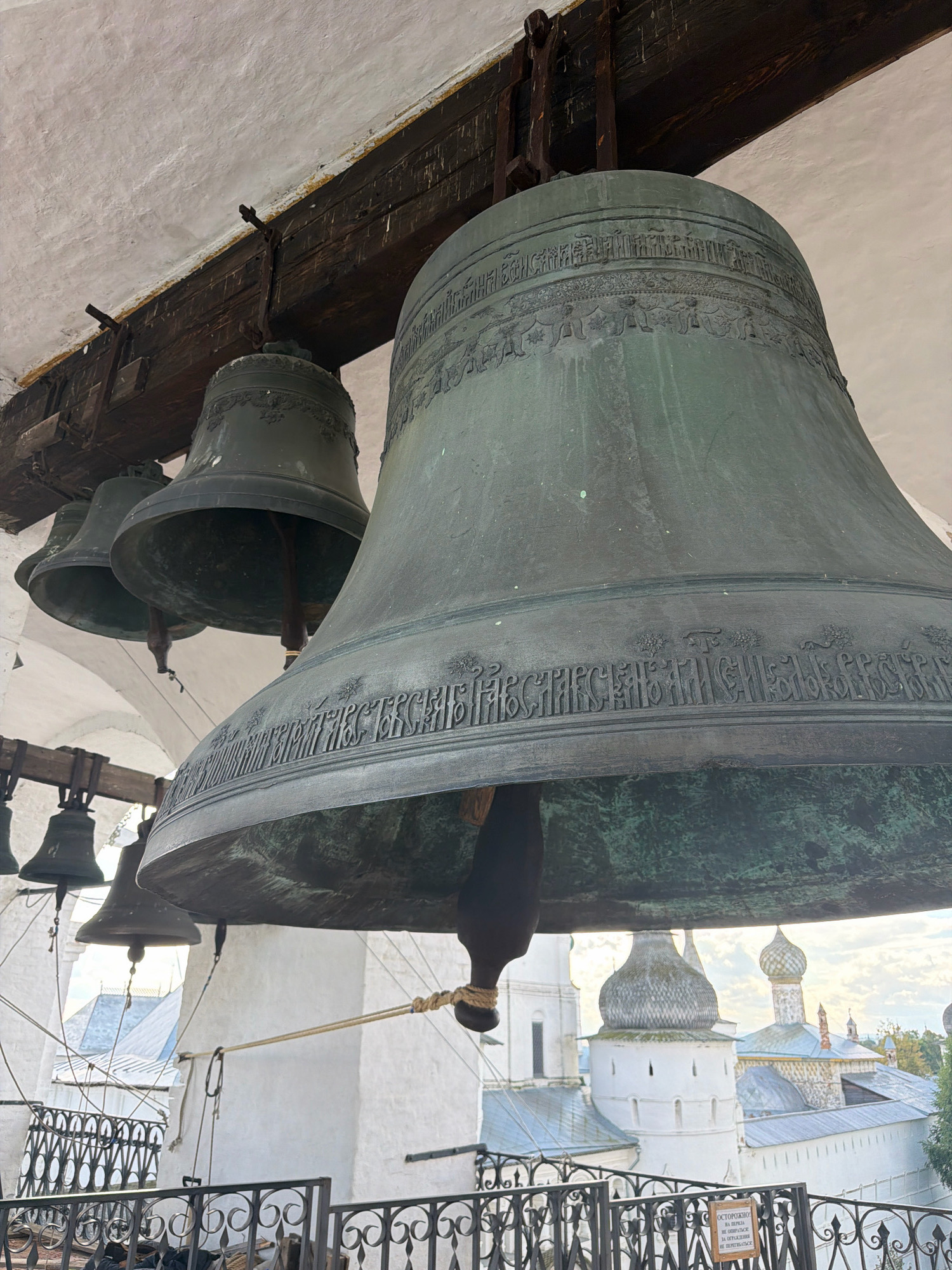 The image shows a close-up view of large, old bells hanging from wooden beams within a bell tower. The bells are made of a dark metal, likely bronze, and are decorated with intricate patterns and inscriptions. In the background, through the open structure of the tower, glimpses of white buildings with domed roofs can be seen, indicative of a Russian Orthodox church or monastery complex. The sky is overcast. There are no people visible in the image. The overall impression is one of historical architecture and religious significance. The location is identified as Ново-Никольское, Russia.