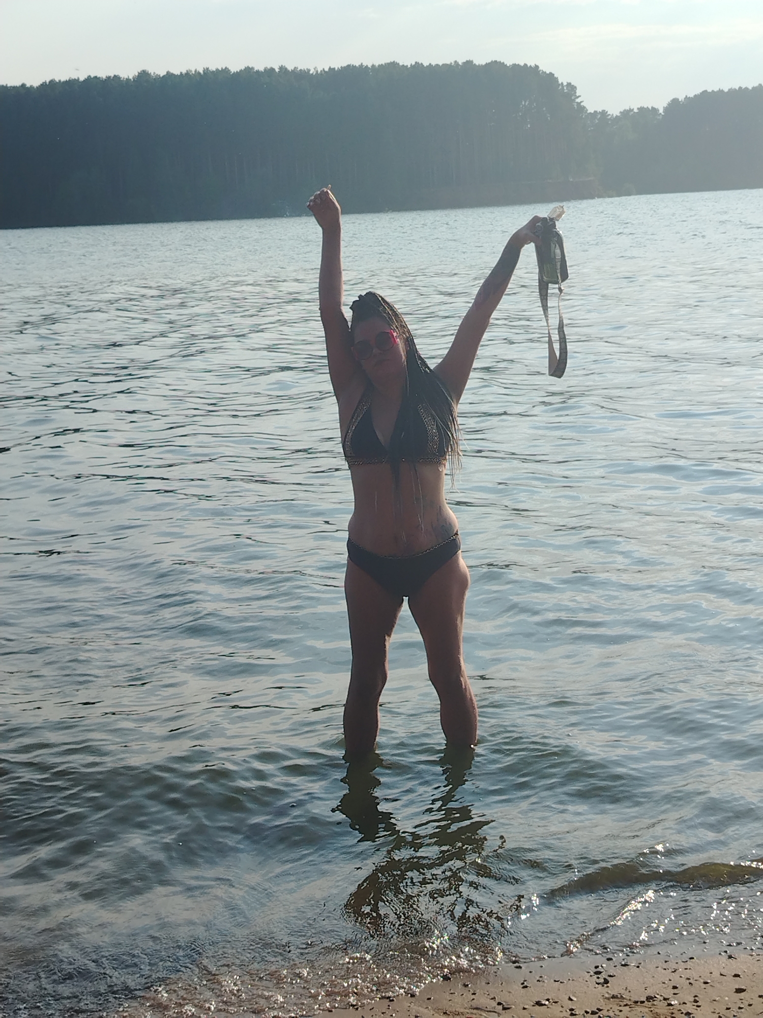 A full-body, medium shot captures a woman standing in the shallow, rippling water of a lake. She is positioned in the center of the frame, facing forward with her arms raised. In her right hand, she holds a dark-colored item, possibly a piece of clothing, with a strap. She wears a dark bikini, sunglasses, and has her hair styled in braids that reach her shoulders. The background displays a tranquil body of water stretching towards a tree-lined shore under a hazy sky. The location is the Kama River bank in Chaikovsky, Russia, characterized by its vast expanse of water and dense forestation typical of the region.