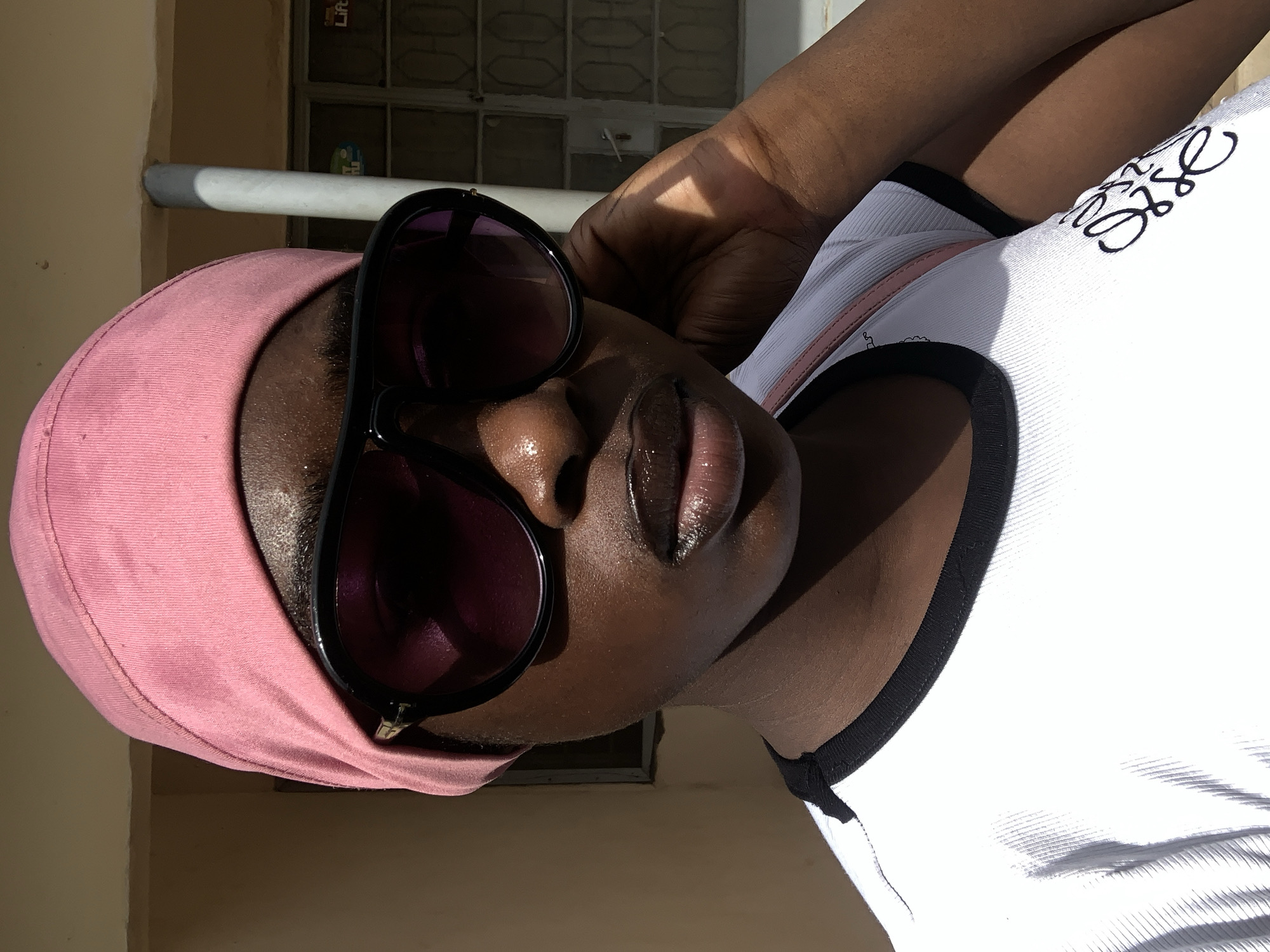A medium shot, rotated 90 degrees clockwise, features a dark-skinned person wearing a pink head covering and dark sunglasses. The person is wearing a white shirt with black trim and an illegible wordmark in cursive on the right side. The person's right arm and hand are raised and visible. A window frame and exterior wall are visible in the background. The location is potentially Rafin Par, Nigeria.
