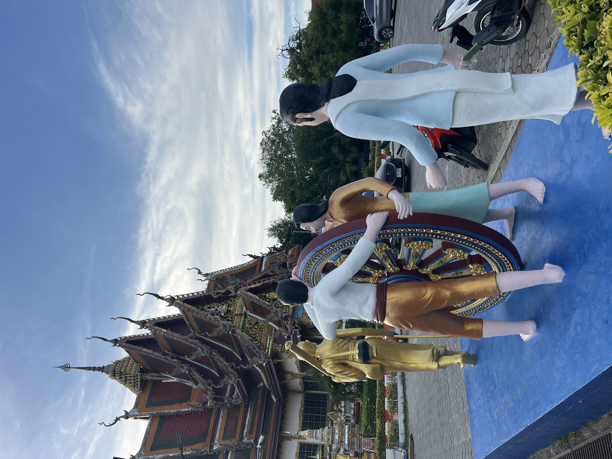 In Baan Plai Laem, Thailand, a religious temple structure with ornate roofs is visible under a cloudy blue sky. In the foreground, a group of colorful statues are arranged on a raised blue platform. Three figures are depicted interacting with a large, ornate wheel, which appears to be a Dharma wheel, a Buddhist symbol. One statue, clad in gold and white, stands to the left, facing away from the viewer. Two other statues, one in gold and light green, and another in white and light blue, are positioned in front of the wheel, seemingly pushing or turning it. A golden statue of a monk is also present, facing the wheel. The scene takes place outdoors, with trees and parked vehicles, including motorcycles, visible in the background.