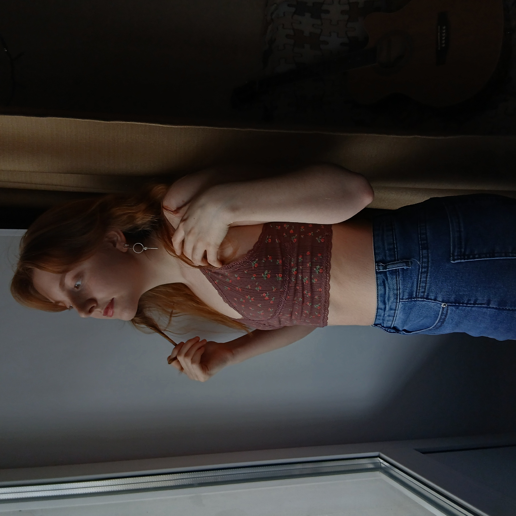 A young, light-skinned woman with red hair is sitting on a light brown surface, possibly a bed or a couch. She is wearing a brown floral patterned bralette and blue jeans. She has a simple earring in her left ear. She's holding a strand of her hair in her right hand. In the background, there is a dark brown guitar and a puzzle. The scene is illuminated by light coming from a window. The specific location in Yekaterinburg, Russia, cannot be determined from the image.