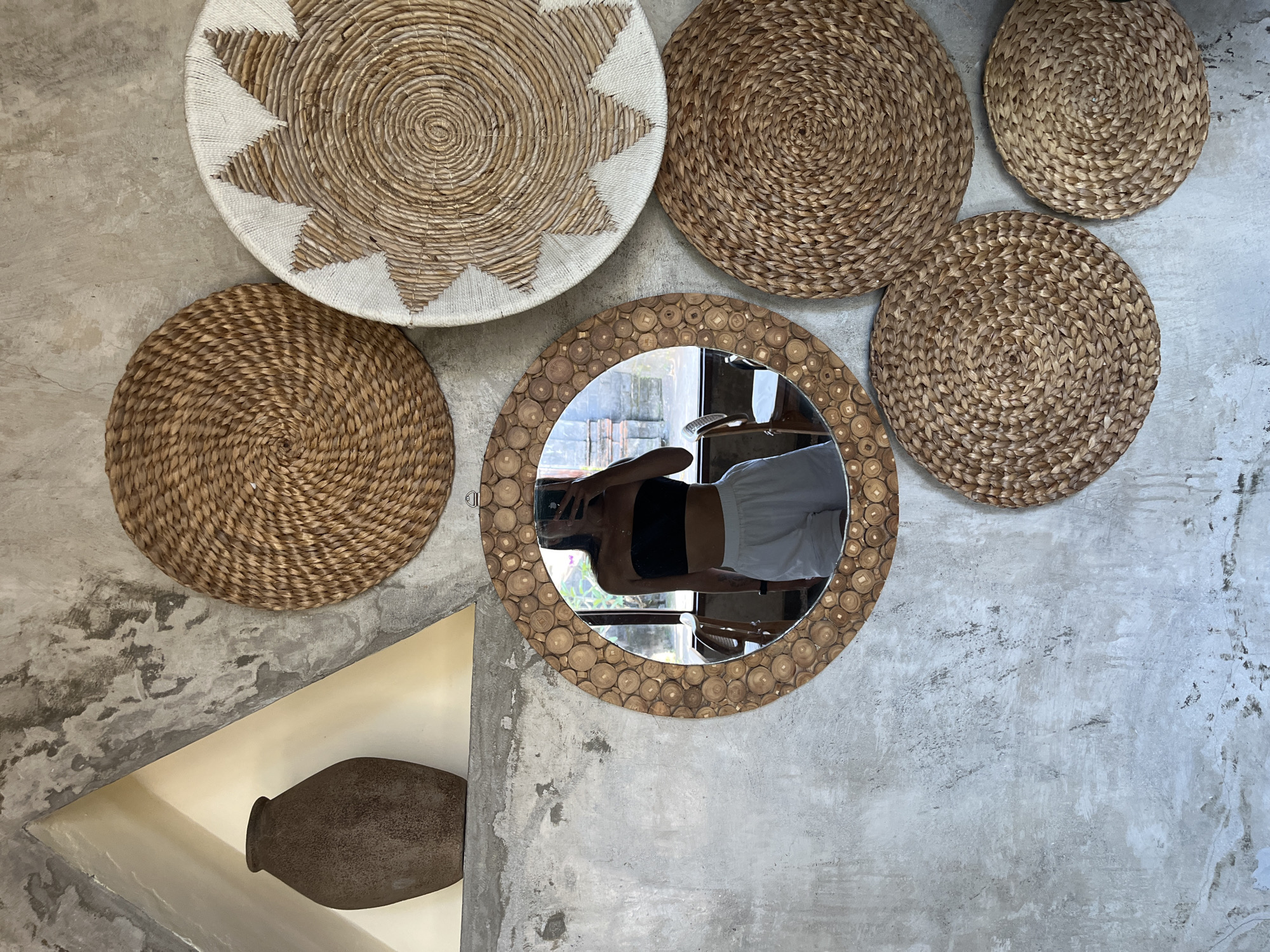 The image depicts a wall decorated with woven circular wall hangings and a mirror, likely in Sukawati, Indonesia, considering the objects and style. A large woven, off-white, and brown disk is positioned at the top left, and several smaller, similarly woven disks of brown, are grouped around it. A circular mirror with a wooden frame made of small wooden pieces is present in the lower center. The mirror reflects a person wearing a black top and white shorts, holding a smartphone. To the left of the mirror, a clay vase rests in a triangular alcove cut into the gray wall. The wall itself has a textured, concrete-like appearance.