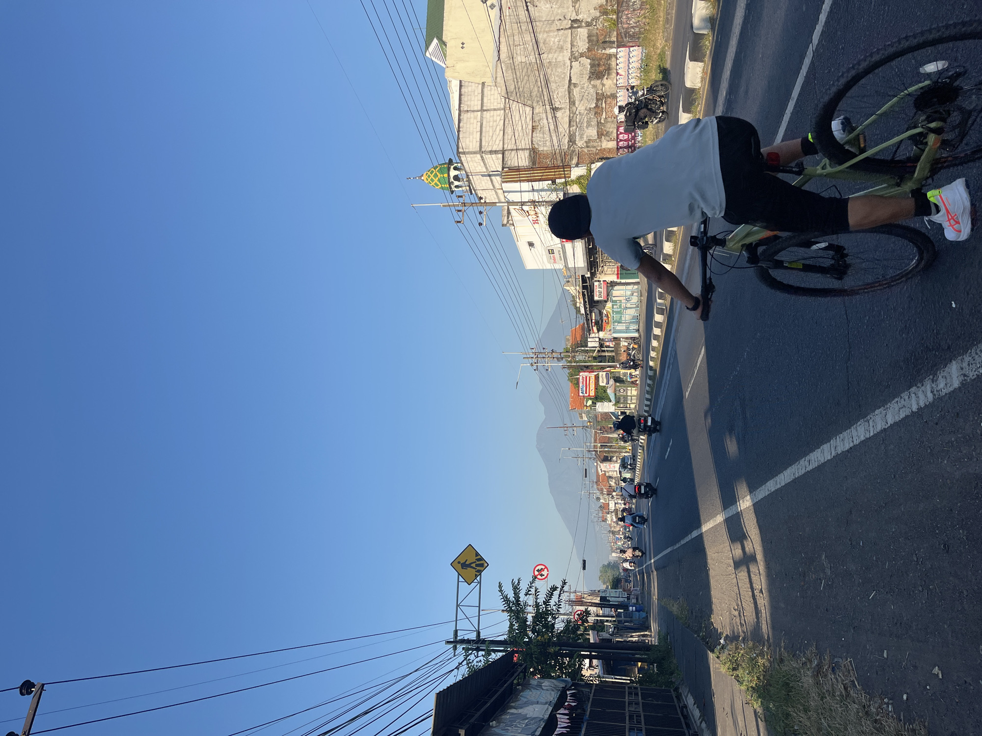A cyclist is riding a bicycle on a road in Siring, Indonesia. The cyclist is wearing a light blue t-shirt, black shorts, and white athletic shoes. The bicycle is green and has black tires. The cyclist is in the foreground, facing away from the camera, and is in the process of pedaling. The road is paved and has a white line dividing the lanes. On the left side of the road, there is a sidewalk with some vegetation and buildings. On the right side of the road, there are more buildings and a mosque with a green and gold dome. In the background, there are mountains visible under a clear blue sky. There are several motorcycles on the road, and some traffic signs. The time of day appears to be morning or late afternoon due to the low angle of the sun.