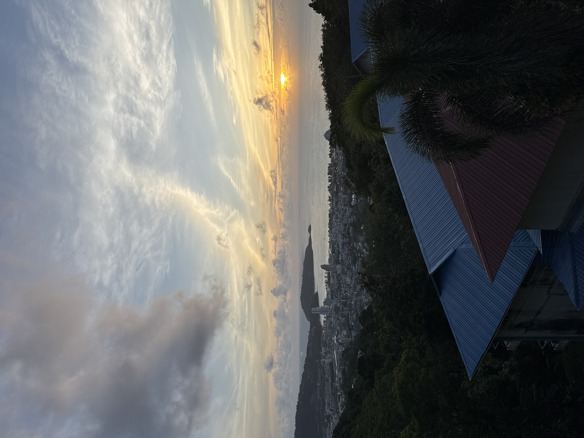 A wide-angle, high-angle shot captures a picturesque sunset over Patong, Thailand. The sky is a canvas of soft hues, with pastel oranges, yellows, and blues dominating the horizon, transitioning to a muted grey and white in the upper portions, accentuated by wispy clouds. The sun, a vibrant orange orb, is partially obscured by a cloud bank at the far right of the frame, casting a warm glow over the tranquil sea. Below the sky, a dense cityscape stretches along the coastline, characterized by numerous buildings of varying heights and architectural styles, interspersed with patches of lush green foliage. In the foreground, the angled rooftops of two buildings, one a metallic blue and the other a rusty red, are visible, partially framing the view. A palm tree frond extends into the frame from the right, adding a touch of tropical greenery. The overall scene depicts a serene evening in Patong, with the city bathed in the gentle light of the setting sun.