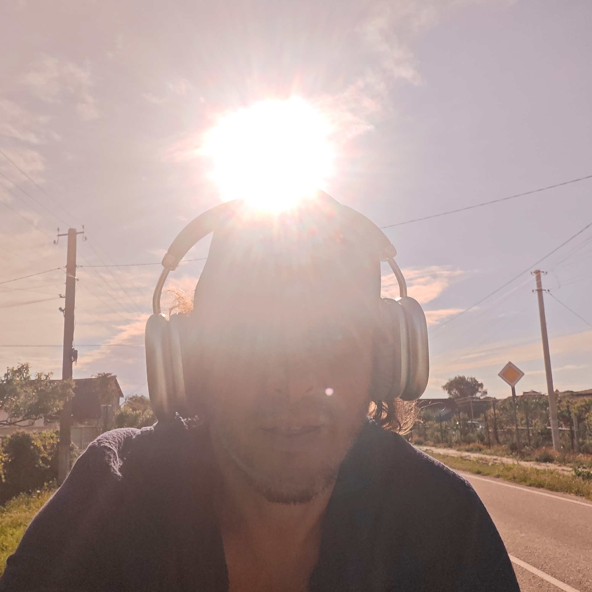 A person wearing headphones is standing on a road in Мысовское сельское поселение, Ukraine. The sun is directly behind the person's head, creating a bright glare. Electrical poles are visible on both sides of the road. A road sign is visible on the right side of the road. Buildings and vegetation can be seen in the background. The person is wearing a dark shirt.