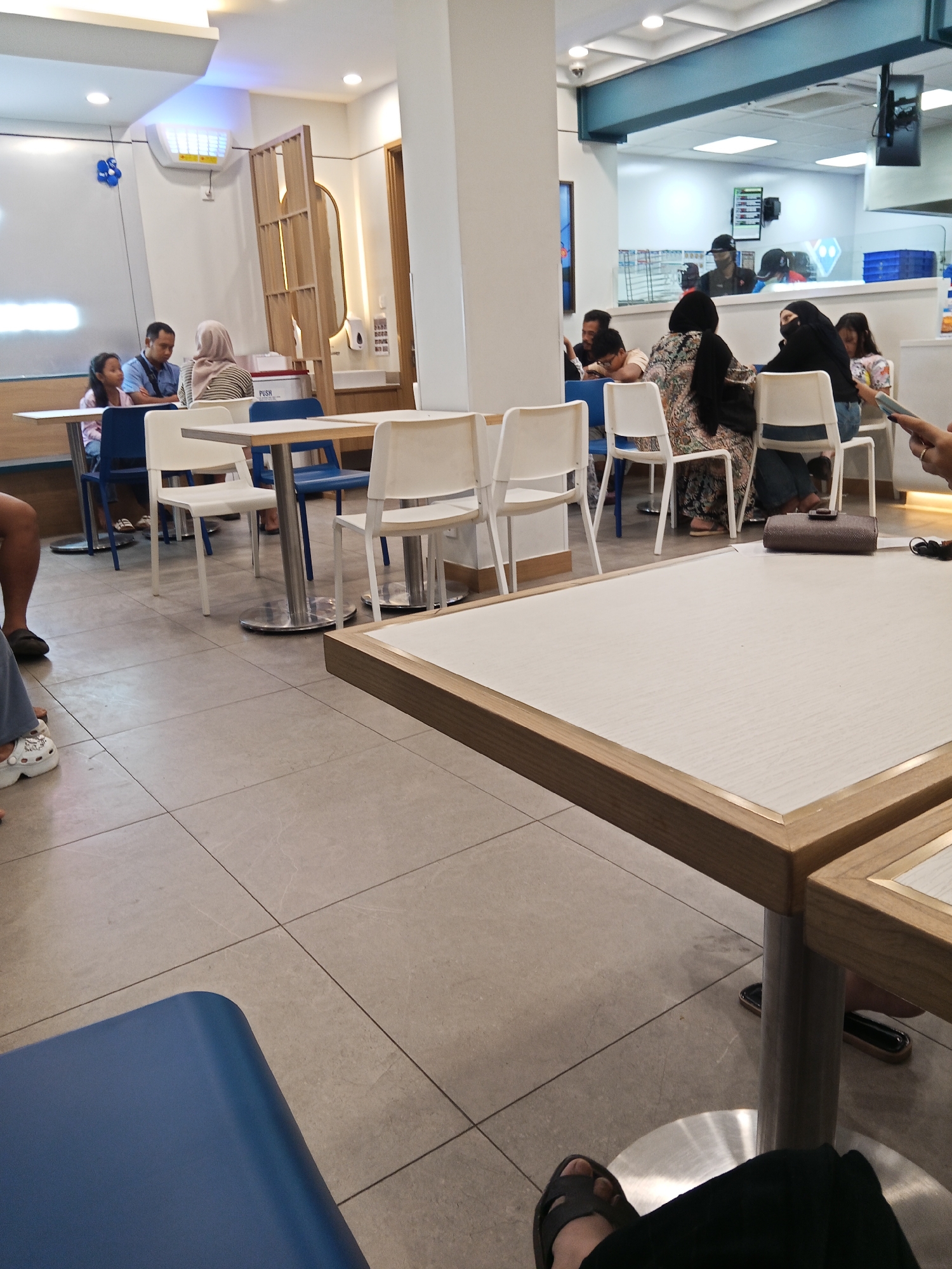 The image depicts the interior of a fast-food restaurant, likely a Domino's Pizza branch given the branding visible in the background. The scene is set in Lubuklinggau, Indonesia. In the foreground, a person's legs and feet are visible, seated at a table. The table has a light-colored surface with a wooden trim. In the mid-ground, several tables and chairs are arranged. Some tables are occupied by customers. On the left, a family group including a man, a woman wearing a hijab, and a young girl are seated. Further back, other customers are seen, including a woman in a hijab and another in a niqab, seated at a table. The restaurant environment appears clean and modern, with tiled flooring and bright overhead lighting. In the background, the service counter area is visible, with employees behind it. The overall atmosphere suggests a casual dining experience.