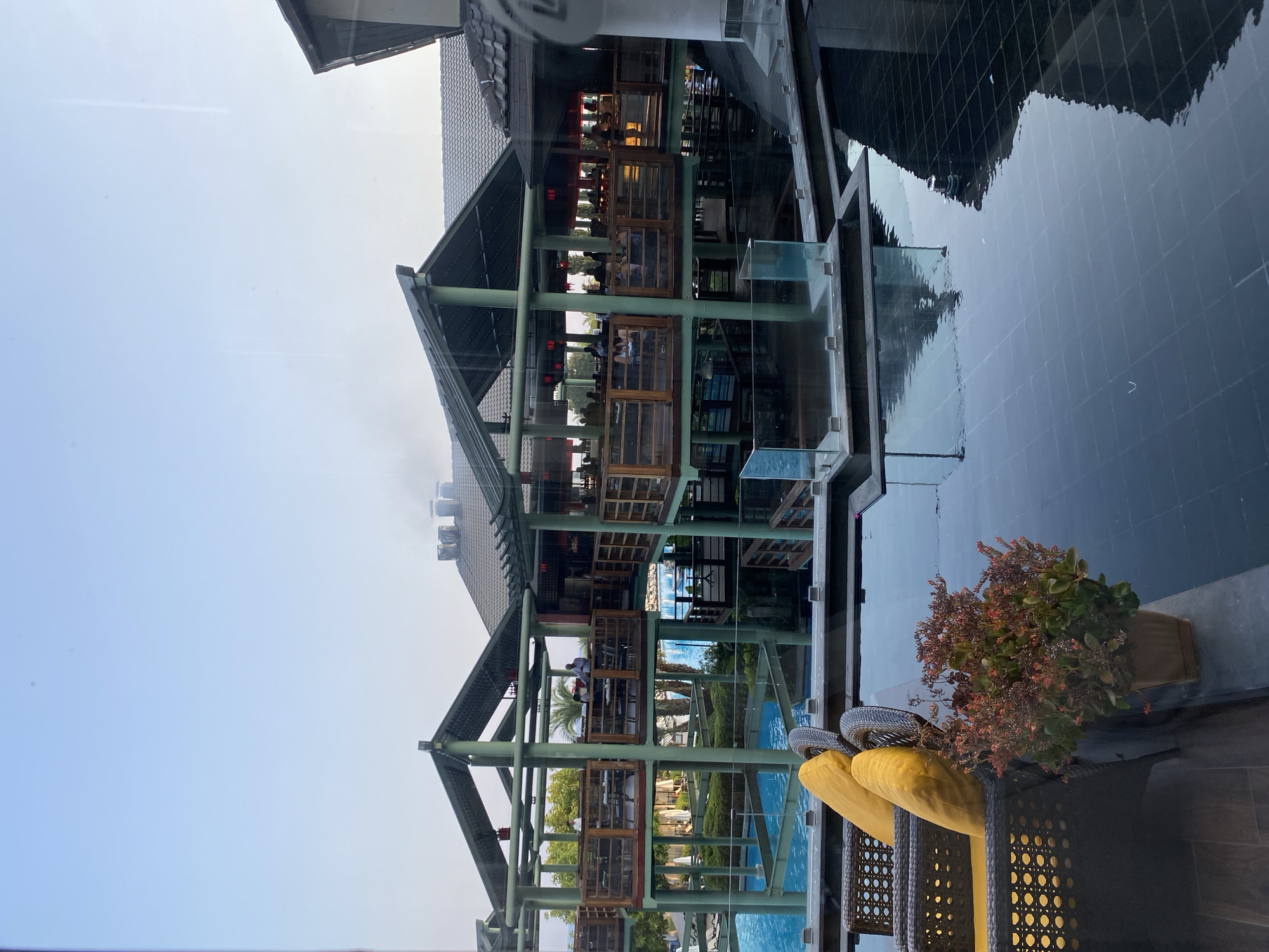 A wide shot captures a resort or hotel complex in Aksu, Turkey, with a multi-level open-air structure resembling a restaurant or dining area overlooking a blue swimming pool. The architecture features a traditional Asian-inspired design with dark tiled roofs and wooden balconies supported by green columns. People are visible dining at tables on the upper levels, suggesting a lively atmosphere. In the foreground, a dark tiled pool with reflections of the architecture and sky is present. Two wicker chairs with bright yellow cushions are placed on a patio area next to the pool, and a potted plant with red and green foliage sits on the edge of the pool. The sky is clear and light blue, indicating daytime.