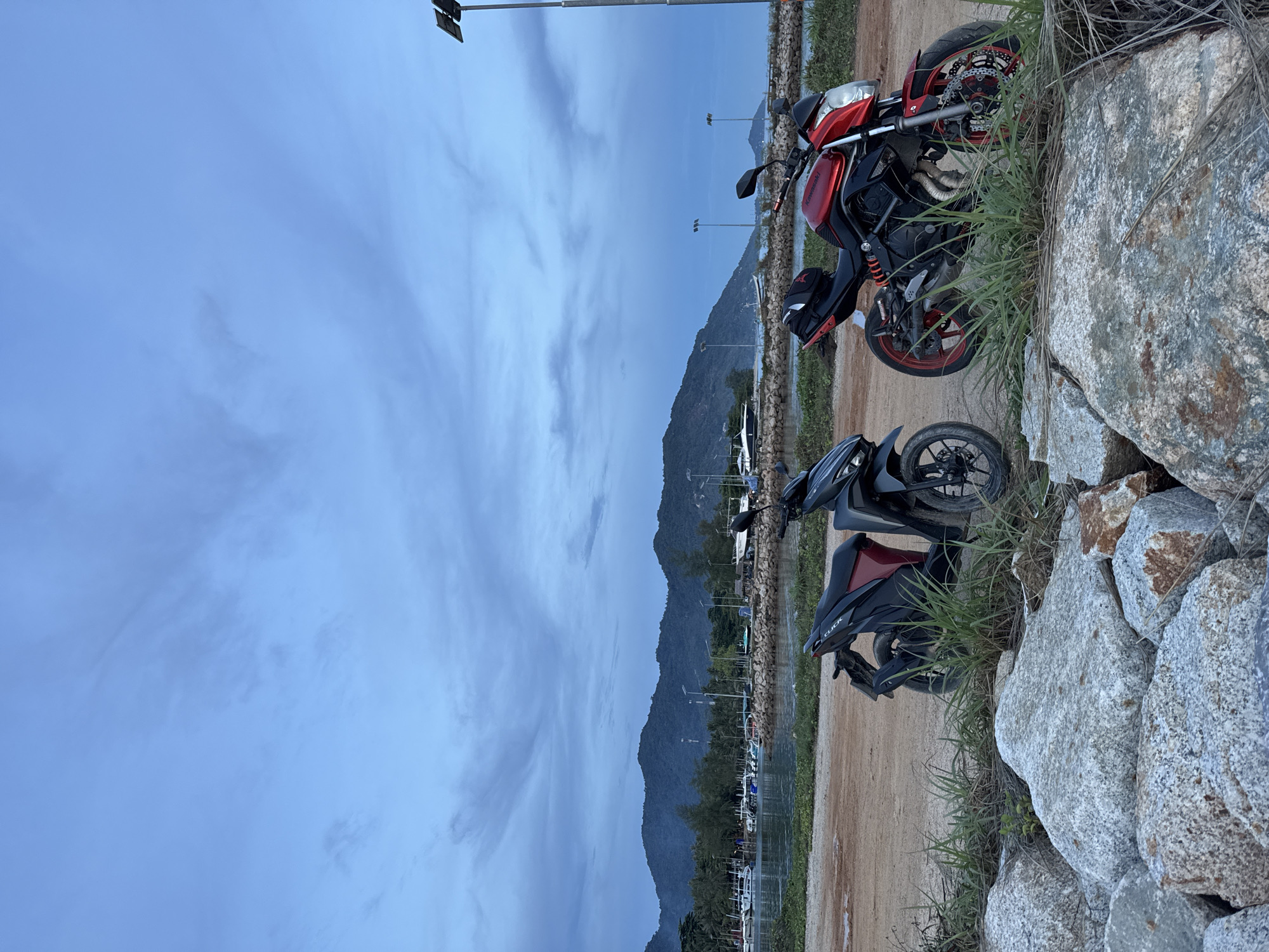 The scene depicts two motorcycles parked along a dirt road adjacent to a rock embankment in Baan Tai, Thailand. One motorcycle is red and black, situated higher on the embankment, partially obscured by grass. The second motorcycle, a black and burgundy scooter, is positioned closer to the road. In the background, a mountain range is visible, with trees lining the base and a strip of boats. Street lights and other vehicles are further in the background. The sky is cloudy.