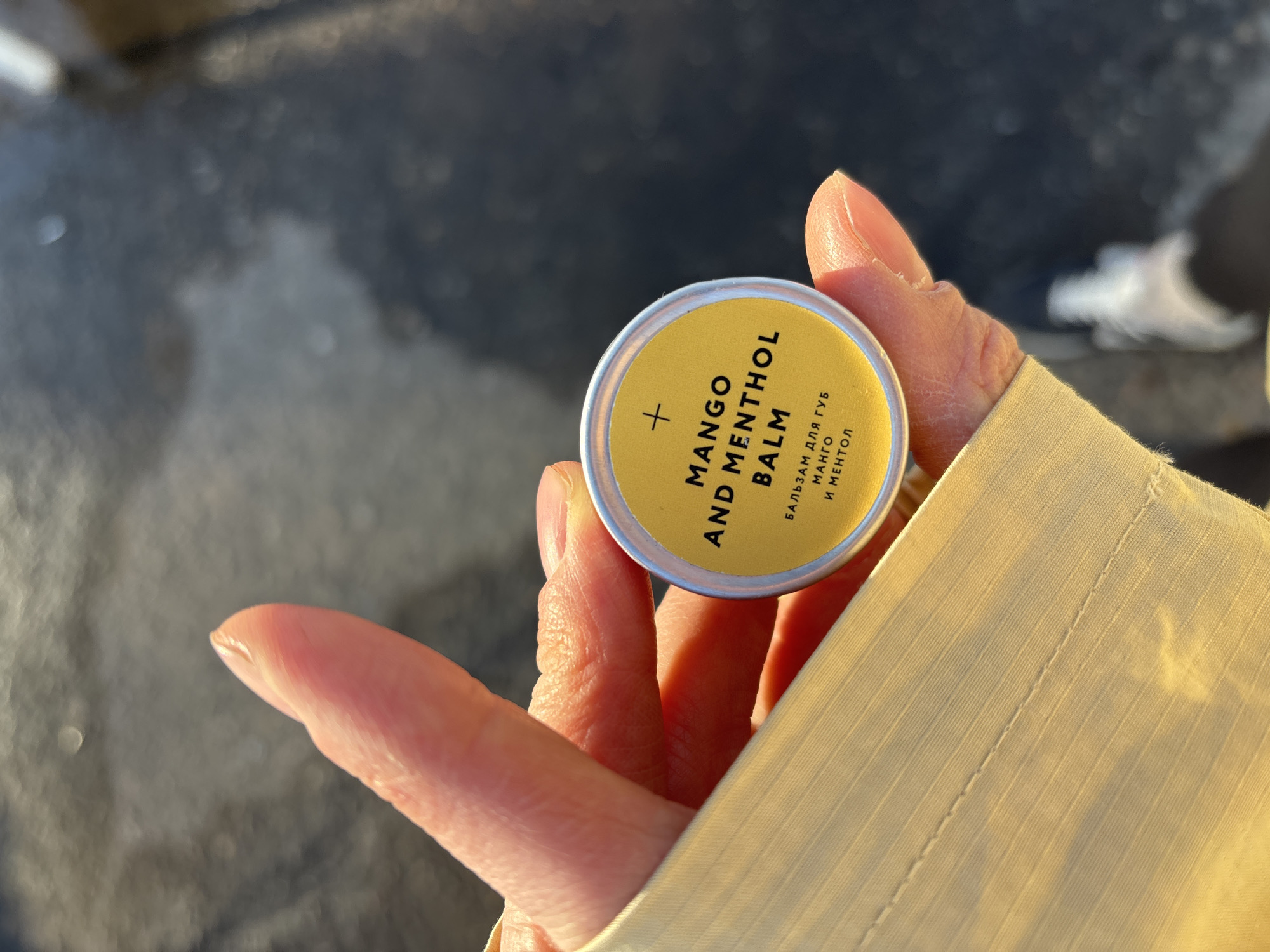 A person is holding a small, round tin of "Mango and Menthol Balm" in their left hand. The hand is fair-skinned and appears to be of a mature individual. The hand is positioned against a background that includes a beige or light yellow fabric sleeve or garment, as well as a blurred out section of a street or ground that seems to have a wet, dark surface. The label on the balm also has text in Russian: "БАЛЬЗАМ ДЛЯ ГУБ МАНГО И МЕНТОЛ." The environment suggests an outdoor urban setting. There is no indication of this being specifically in the city of Chaikovsky, Russia. The focus is on the hand holding the balm and the immediate background.
