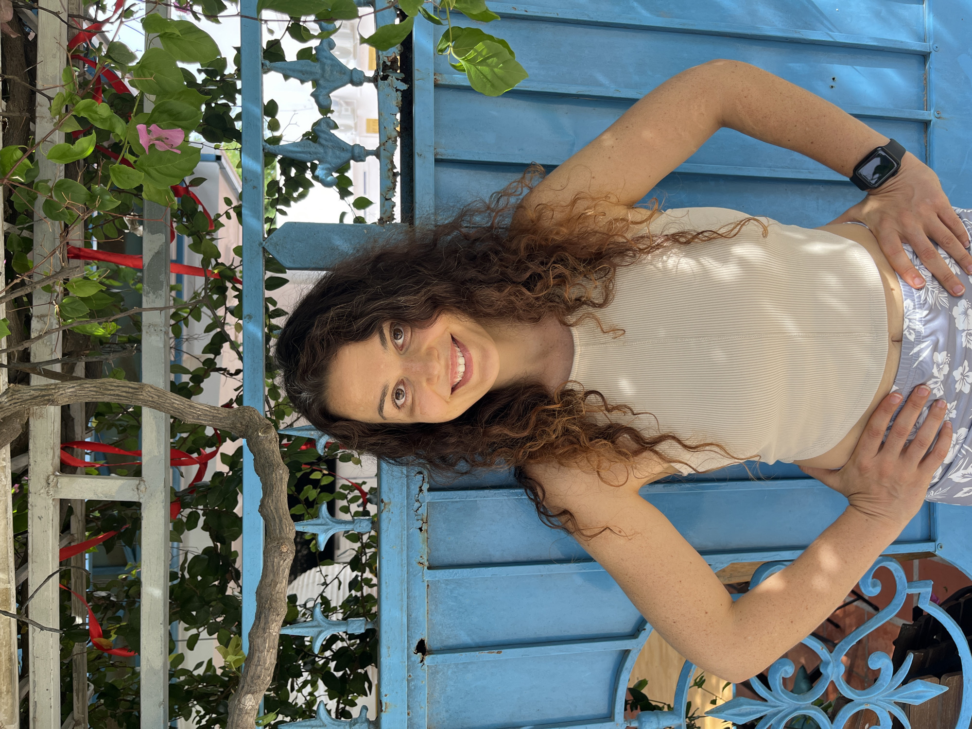 A woman with curly brown hair stands in front of a bright blue metal gate. She is wearing a beige ribbed tank top and light-colored patterned shorts. Her hands are placed on her hips, and she is looking at the camera with a smile. Green foliage and red ribbons are visible above and behind her, suggesting an outdoor setting with some decorative elements. The blue gate appears to be part of a fence or entrance. The lighting suggests it is daytime. The overall scene is casual and outdoors.