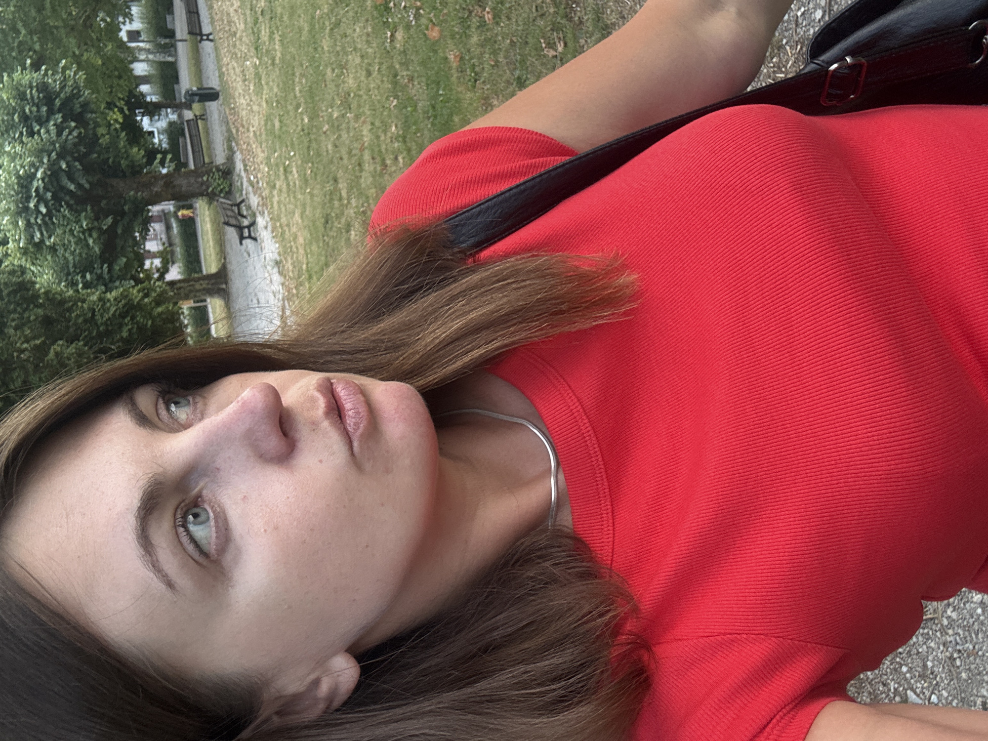 A close-up, profile shot shows a woman with medium-length brown hair and light blue eyes looking to her right. She is wearing a red, ribbed t-shirt and a thin silver necklace. Her left shoulder and a black bag strap are visible. The background is blurred, showing a park with grass, trees, and benches. The scene appears to be in a public park, likely during the daytime. There is no indication of the specific location within Saint-Claude, France.