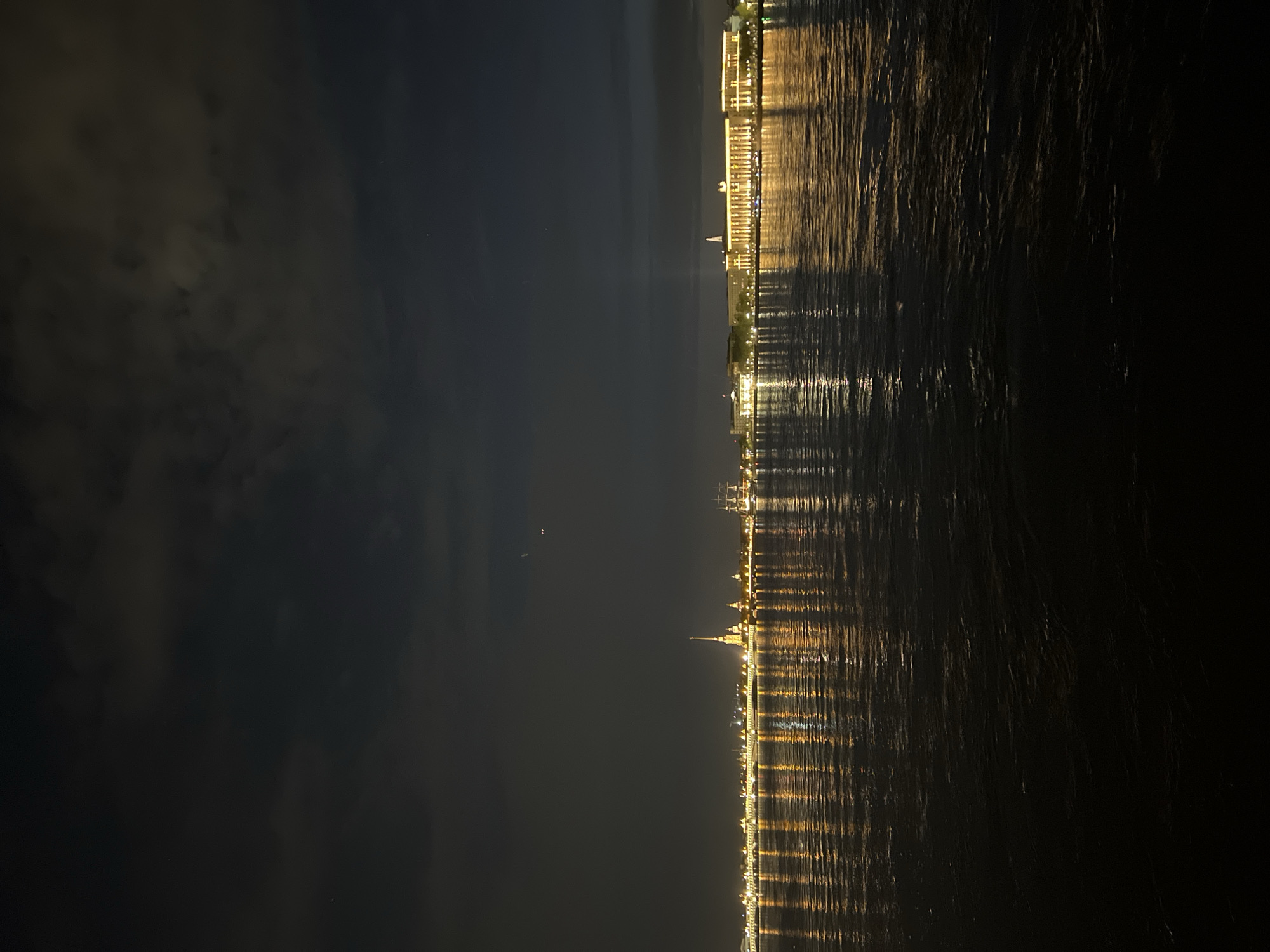 The image shows a nighttime view of Saint Petersburg, Russia. The buildings along the Neva River are illuminated, and their lights are reflected in the water. Notable landmarks include the Peter and Paul Fortress with its distinctive golden spire. The sky is dark, with some clouds visible. The image appears to be taken from across the river, capturing the city skyline.