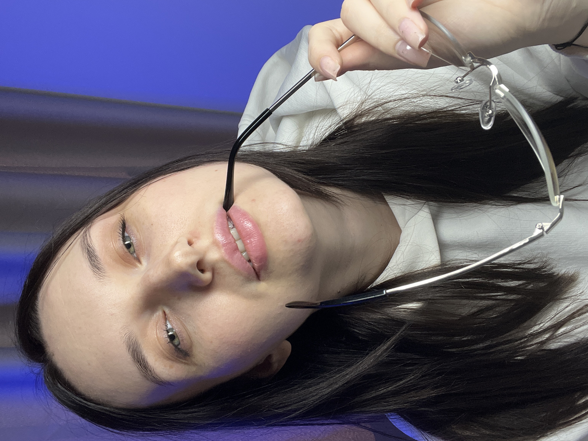 A close-up indoor portrait depicts a young adult female, located in an unspecified indoor setting within the city of Tyumen, Russia. The subject, possessing long dark hair, fair skin, and green eyes, gazes directly into the camera. She is attired in a white hooded garment. She holds a pair of metal-framed eyeglasses; specifically, the black-tipped right temple arm is placed between her slightly parted lips, while the remaining portion of the eyeglasses, including the frame and left temple arm, is held by her left hand. Her left hand's fingernails are visible, covered with a light, possibly clear or nude, polish. The background is out of focus, featuring a dark, vertically draped fabric element on the left side and a vibrant blue illuminated surface on the right. A subtle purple light cast is discernible on the left side of the subject's hair and face, consistent with ambient light interacting with the blue background illumination.