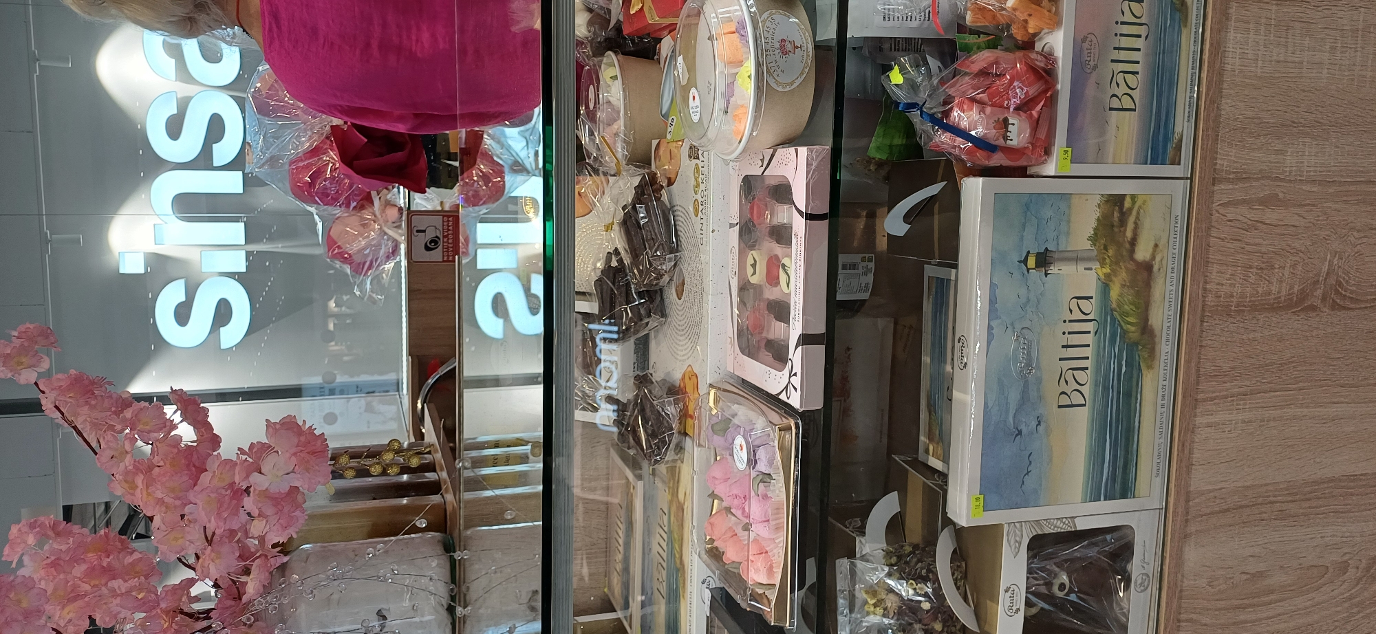 The image shows a display case filled with various chocolates and sweets. In the foreground, there are boxes of chocolates labeled "Baltija" with a lighthouse and sea scene depicted on them. To the left, there's a box containing individually wrapped pink and purple rose-shaped candies. To the right, there is a clear plastic container with small, colorful candies. In the background, a large sign with the word "Sinsay" is visible, illuminated from behind. A person wearing a bright pink top is partially visible on the right side of the image. The display case is made of glass, and the shelves are stocked with packaged confectionery. The overall environment appears to be a retail store or a confectionery shop.