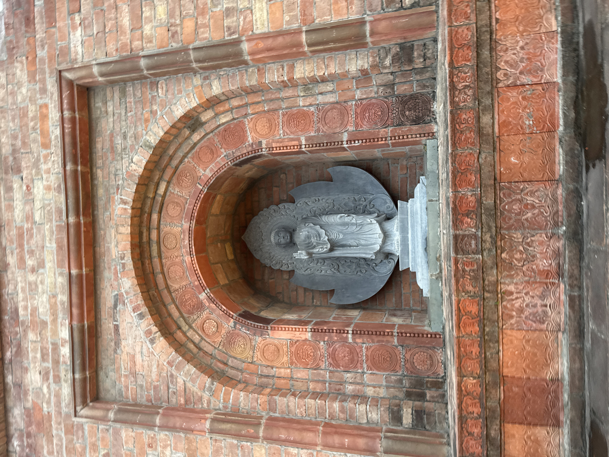 An outdoor, medium shot captures a weathered, arched brick niche containing a gray stone statue of the Buddha. The niche is framed by red brickwork with decorative circular motifs etched into the inner arch and a band of carved rectangular panels below. The Buddha statue stands upright, facing forward, with its right hand raised in a gesture of blessing. Its left hand rests by its side, and it wears traditional monastic robes. A large, fan-shaped gray stone halo is positioned behind the statue. The brickwork appears aged, with some discoloration and signs of wear. The surrounding environment is not visible, but the statue and niche suggest a religious or historical site in Tây Hoa Lư Ward, Vietnam.